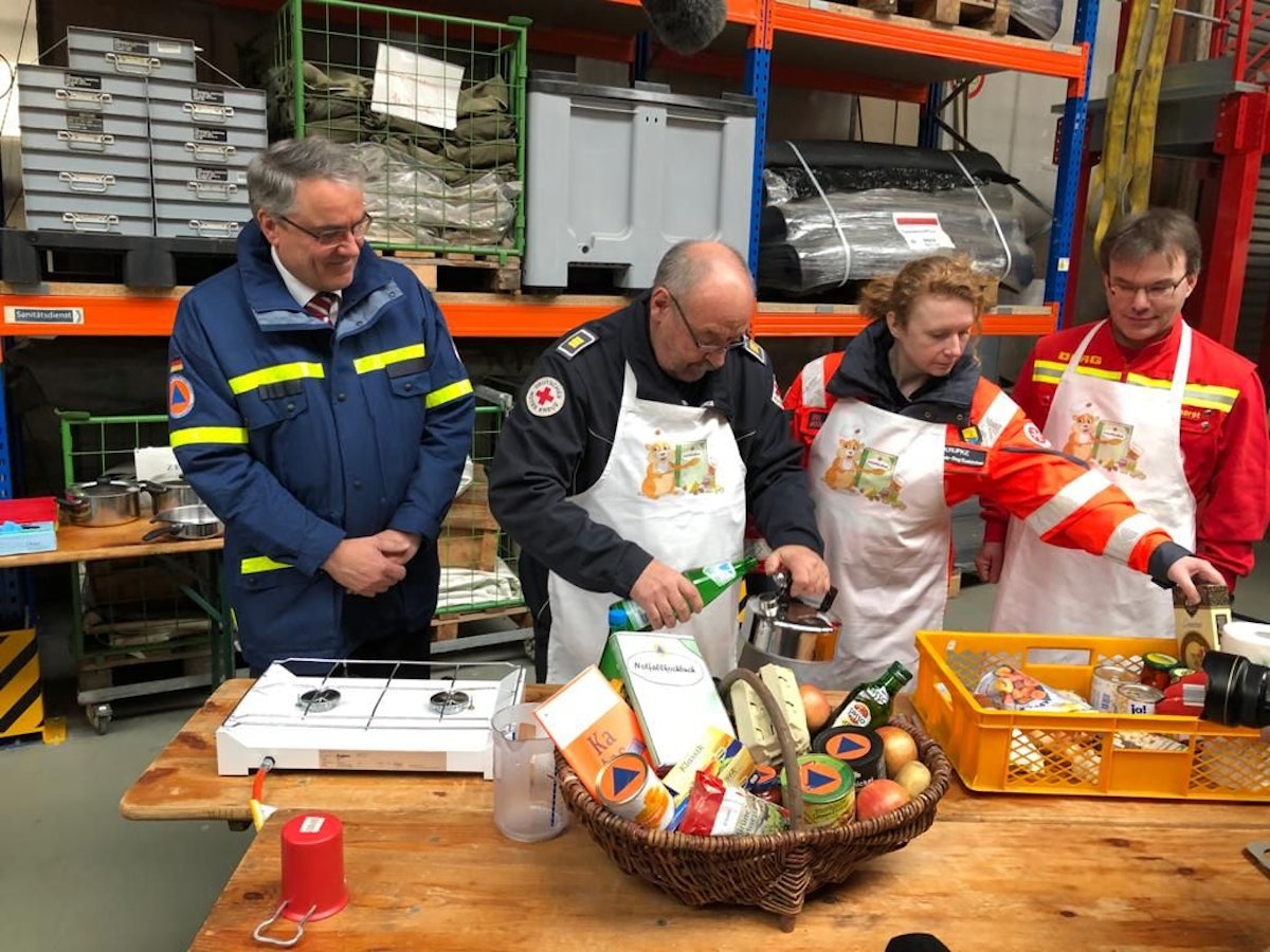 Dr. Thomas Herzog (BBK), Peter Winter vom DRK, Stefanie Krupke (Johanniter) und Thomas Stockhorst (DLRG) stehen an einem Tisch, auf dem sich ein Korb mit Lebensmitteln, frische sowie aus der Dose, befinden.
