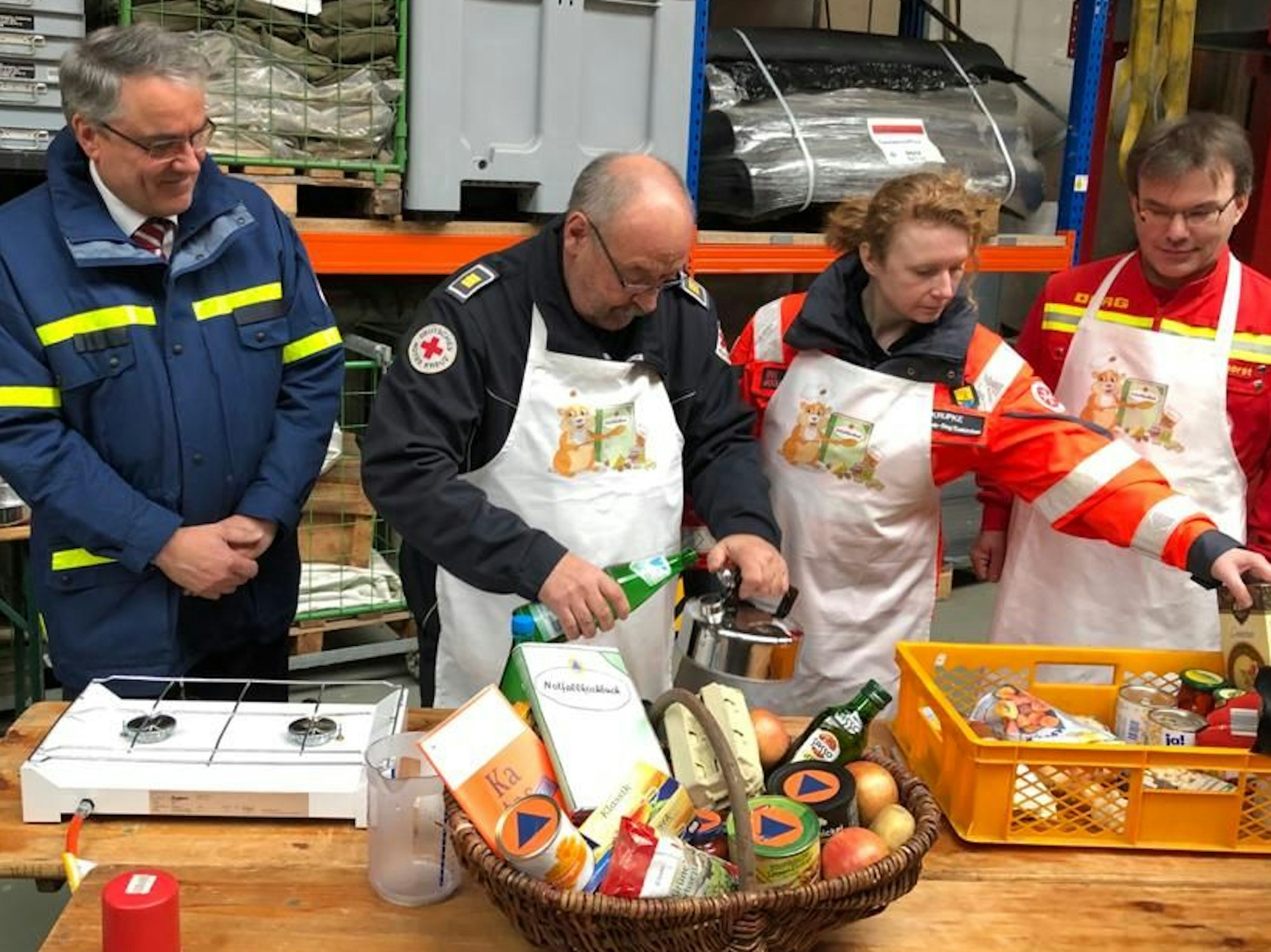Dr. Thomas Herzog (BBK), Peter Winter vom DRK, Stefanie Krupke (Johanniter) und Thomas Stockhorst (DLRG) stehen an einem Tisch, auf dem sich ein Korb mit Lebensmitteln, frische sowie aus der Dose, befinden.