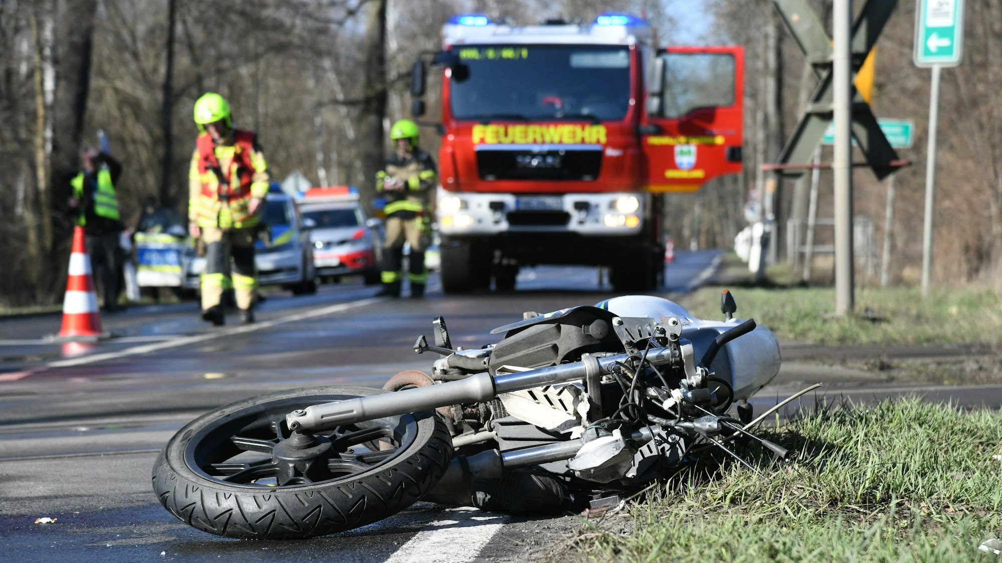 Ein Motorrad liegt an einem Bahnübergang neben der Straße. Polizei und Feuerwehr sind im Hintergrund zu sehen.