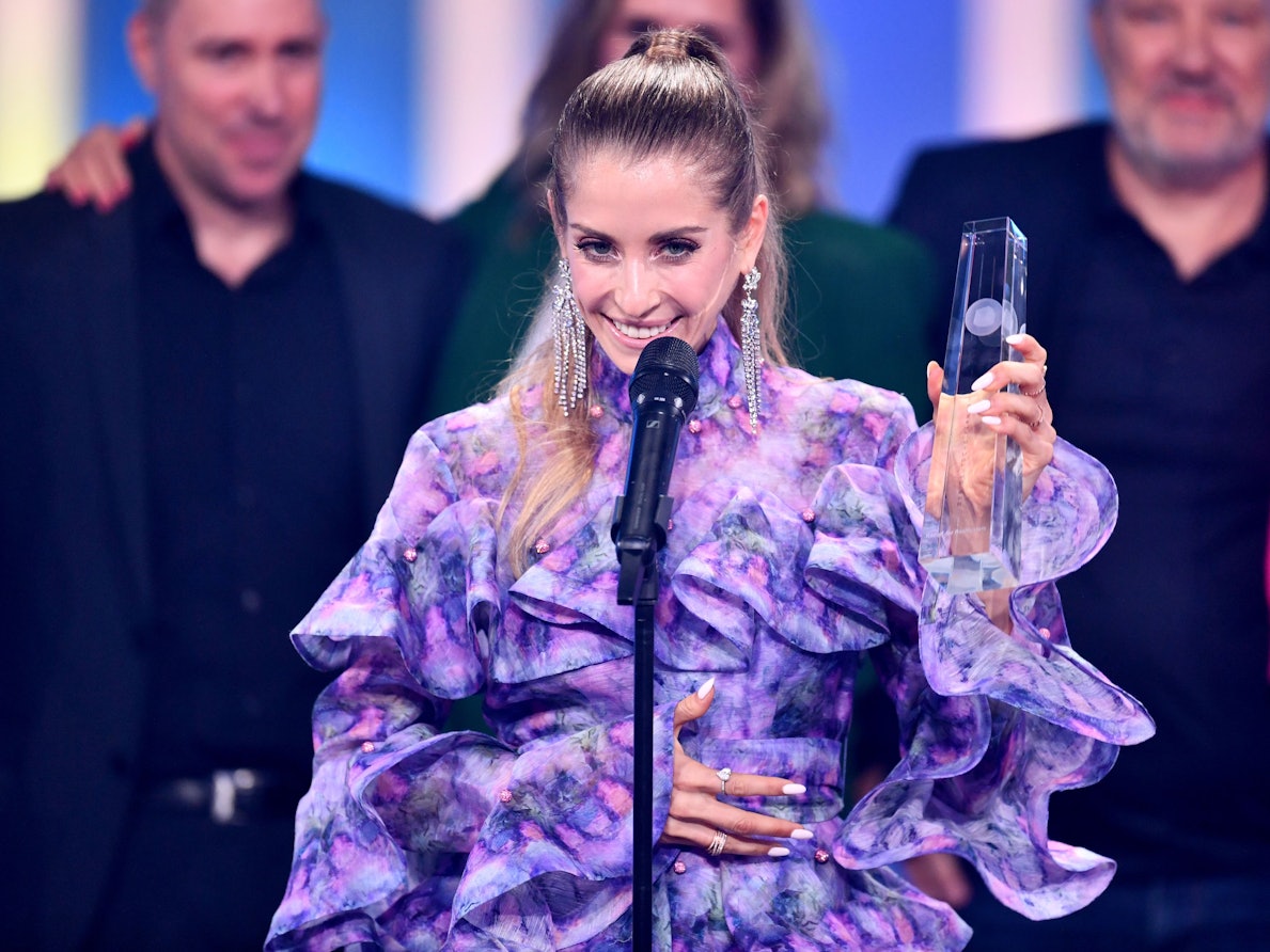 Auf dem Foto steht Cathy Hummels auf der Bühne und nimmt ihre Auszeichnung entgegen. Sie trägt ein lilafarbenes Kleid. Ihre Haare hat sie zu einem strengen Pferdeschwanz zusammengebunden.