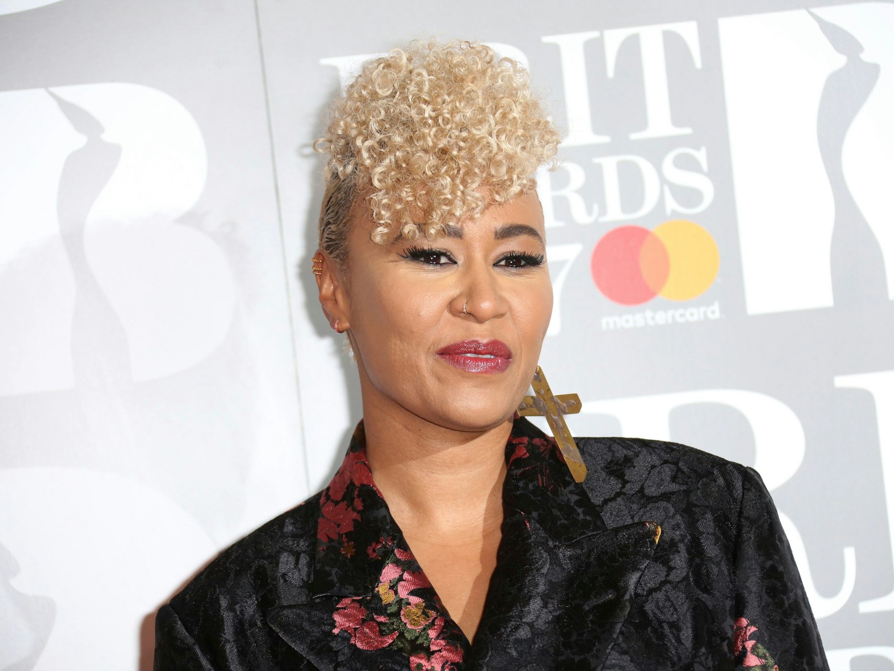 Sängerin Emeli Sandé präsentiert ihren blonden Lockenschopf in einem schwarzen Kleid, das mit Blumenmustern versehen ist. Ein weiterer Hingucker: Am rechten Ohr trägt sie ein Kreuz als Ohrring.