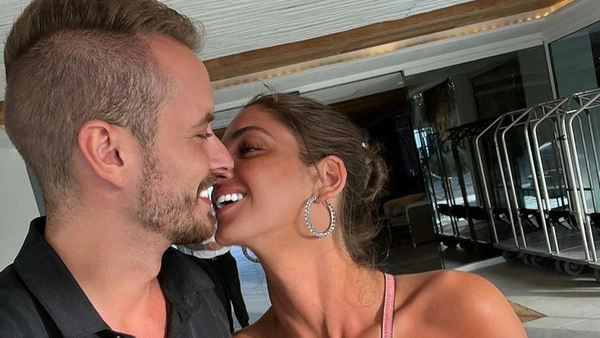 Auf dem Bild schauen sich Julian und Tanja verliebt an. Sie trägt ein trägerloses weißes Top und eine rosafarbene Handtasche. Julian trägt ein dunkelgraues Hemd.