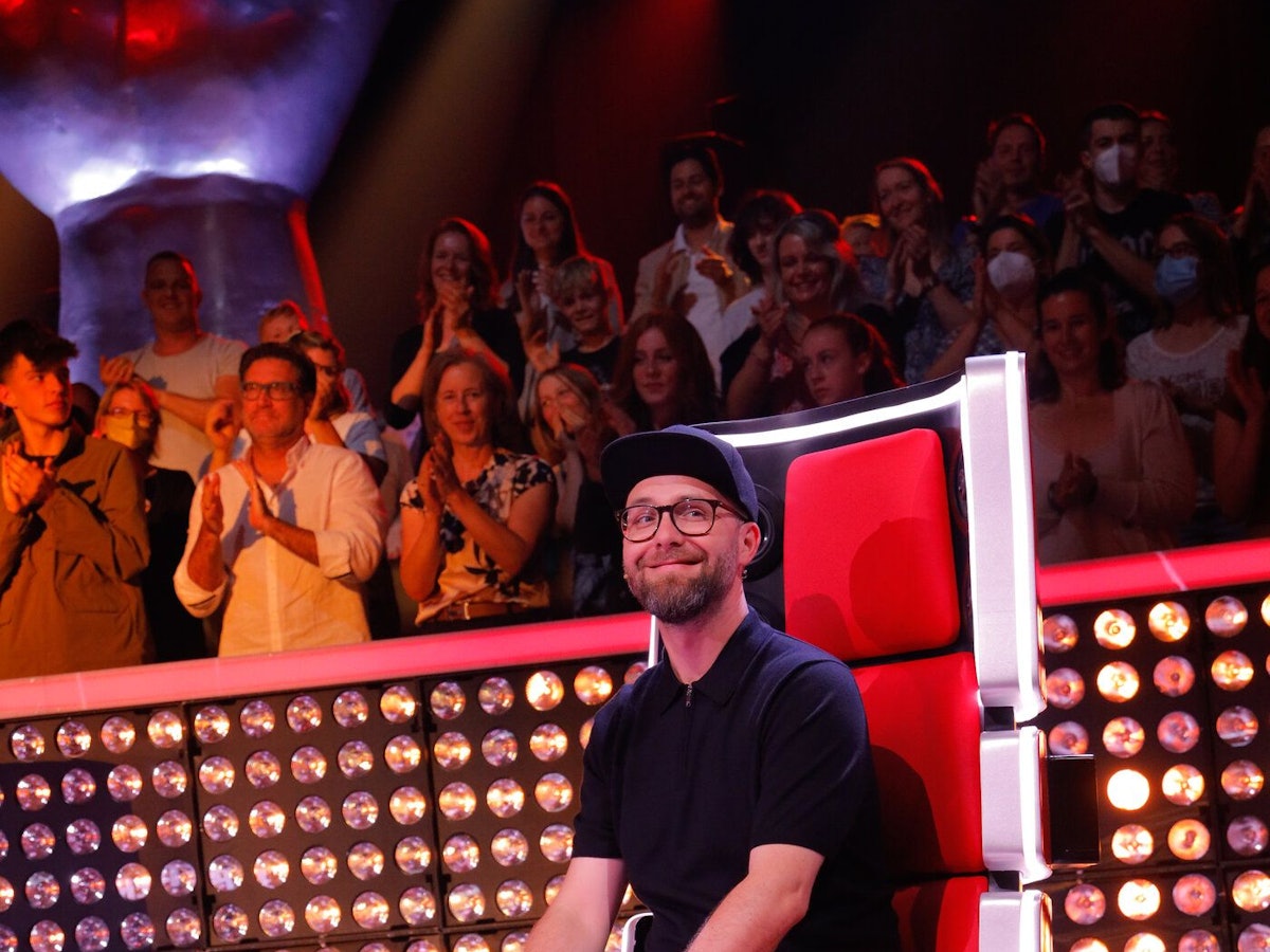 Mark Forster sitzt auf seinem Jury-Stuhl. Er trägt eine helle Hose, ein dunkles Shirt und eine schwarze Kappe.