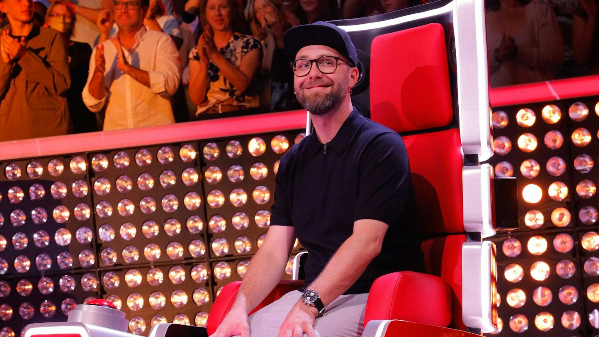 Mark Forster sitzt auf seinem Jury-Stuhl. Er trägt eine helle Hose, ein dunkles Shirt und eine schwarze Kappe.