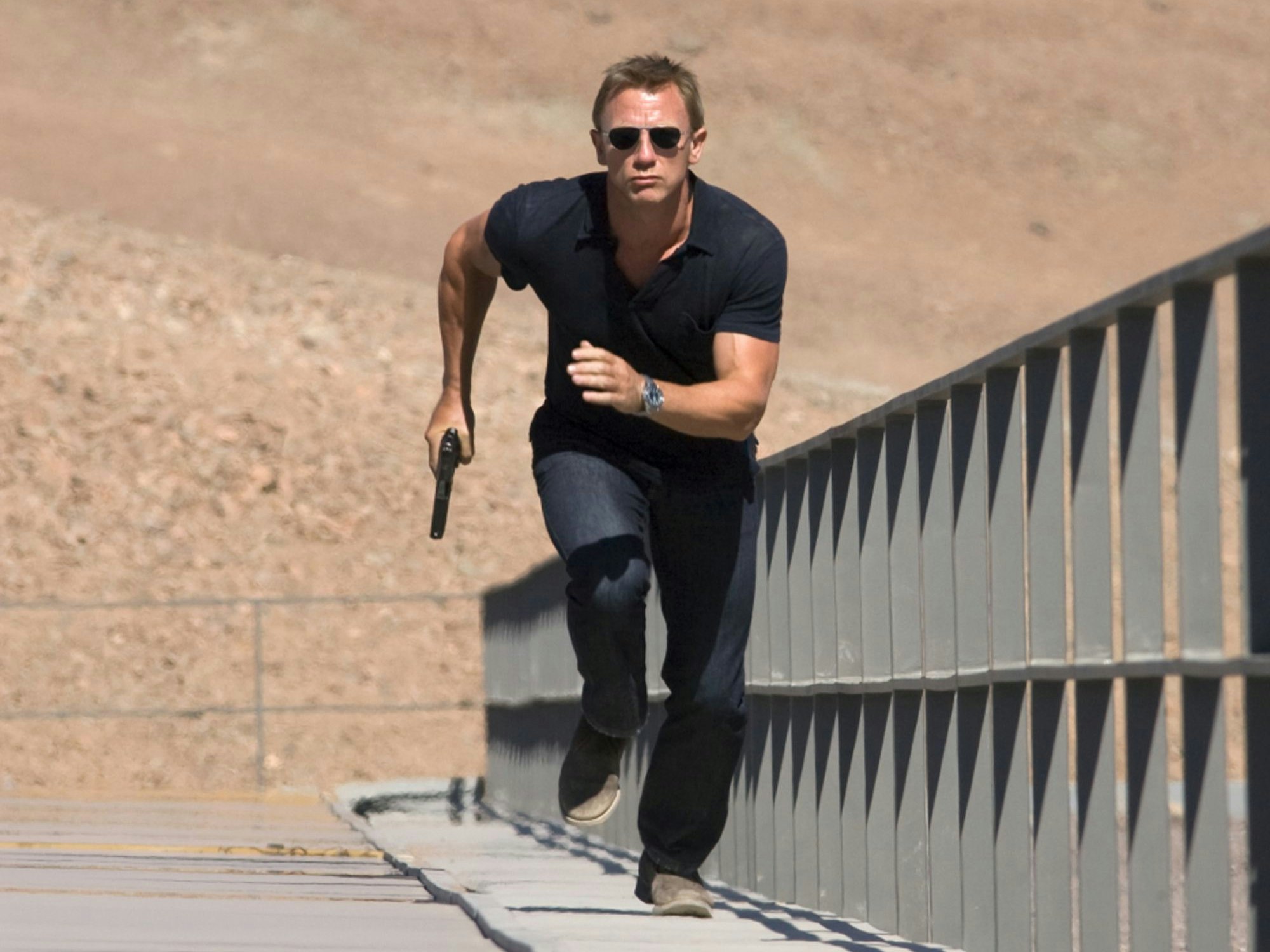 Daniel Craig rennt in der Szene des Films „Ein Quantum Trost“ einen Weg entlang. In der rechten Hand hält er eine Pistole. Er trägt Jeans, ein schwarzes T-Shirt und eine dunkle Sonnenbrille.