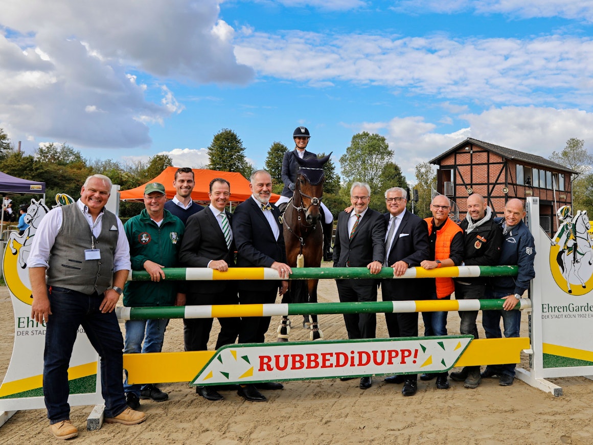 Mehrere Männer stehen an einem Hindernis, hinter ihnen eine Frau zu Pferd.