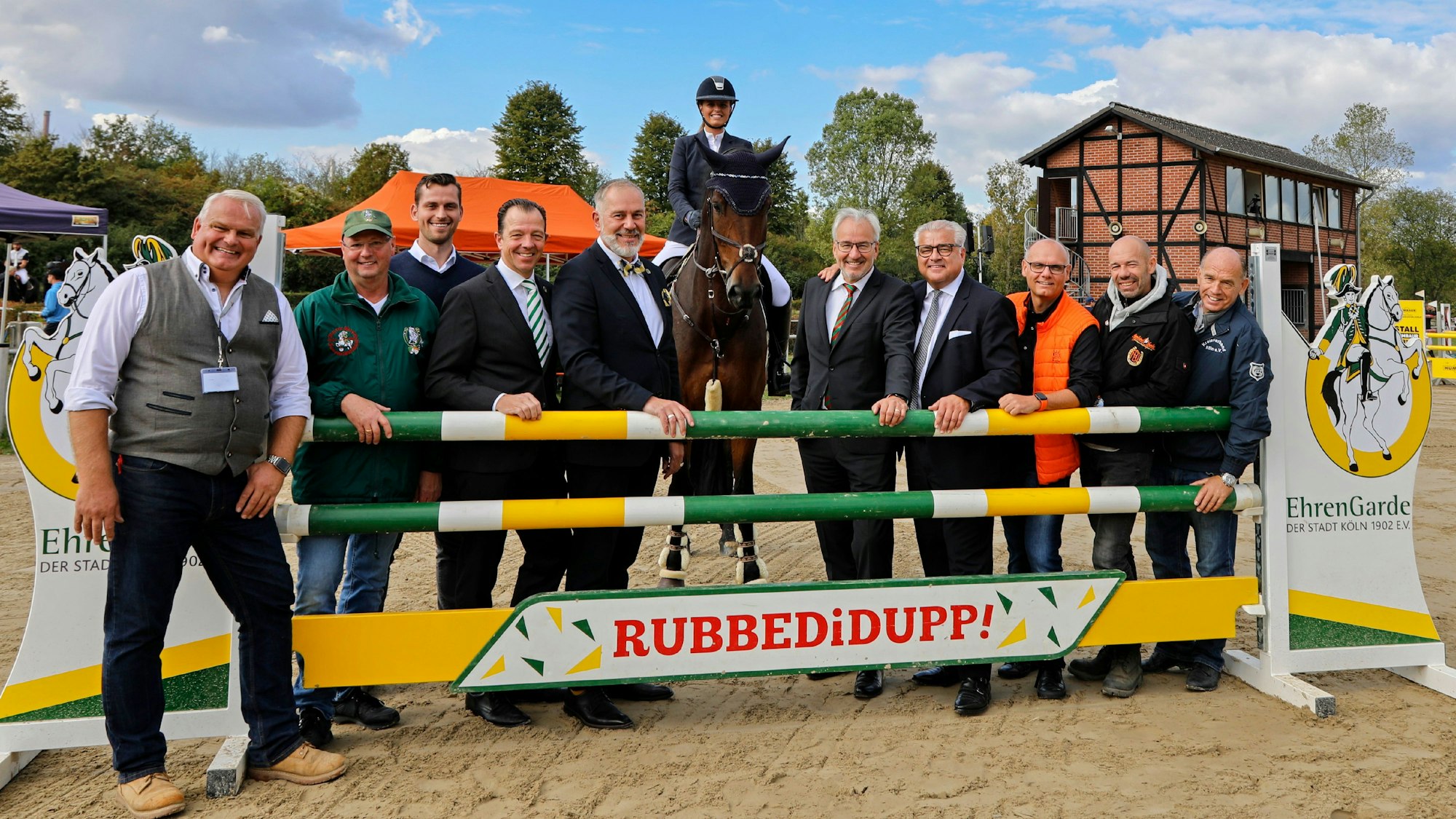 Mehrere Männer stehen an einem Hindernis, hinter ihnen eine Frau zu Pferd.
