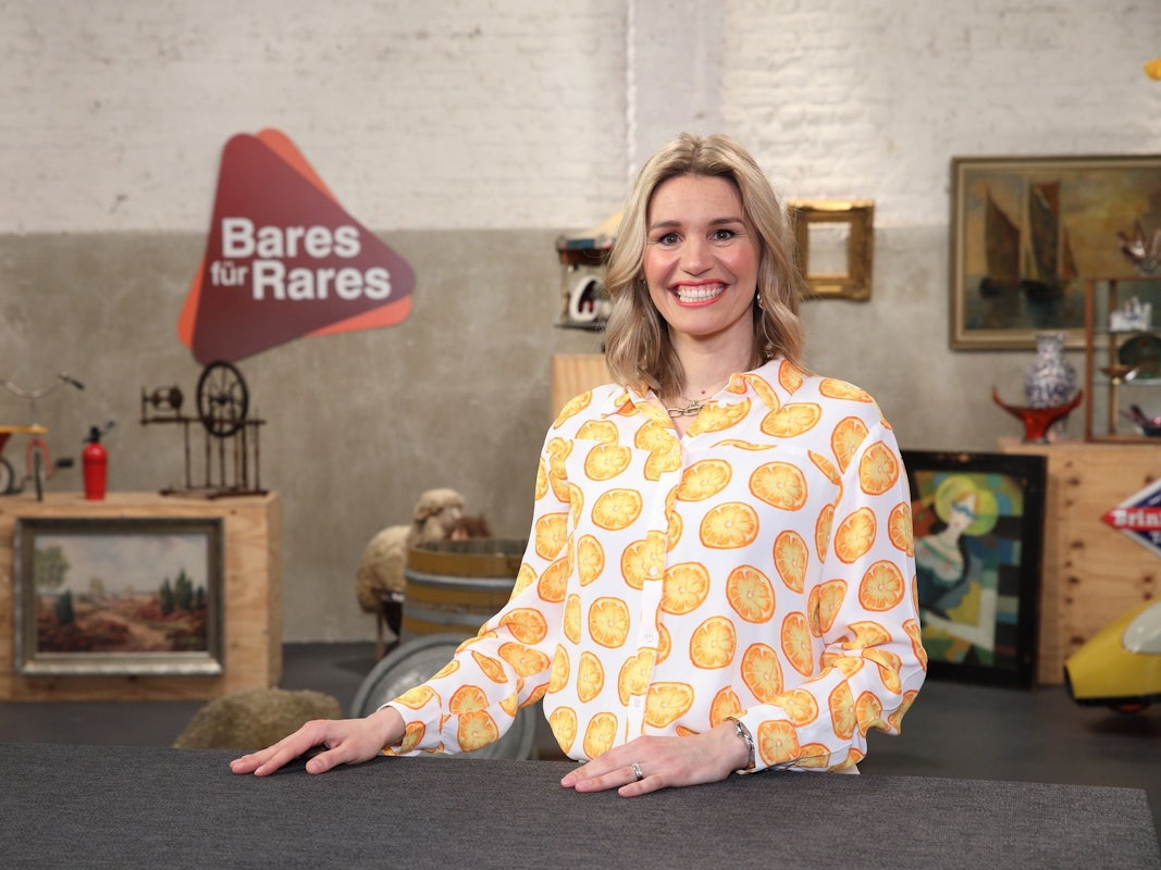 Dr. Bianca Berding steht am Expertentisch und lächelt in die Kamera. Sie trägt eine weiße Bluse mit Orangenscheiben als Muster.