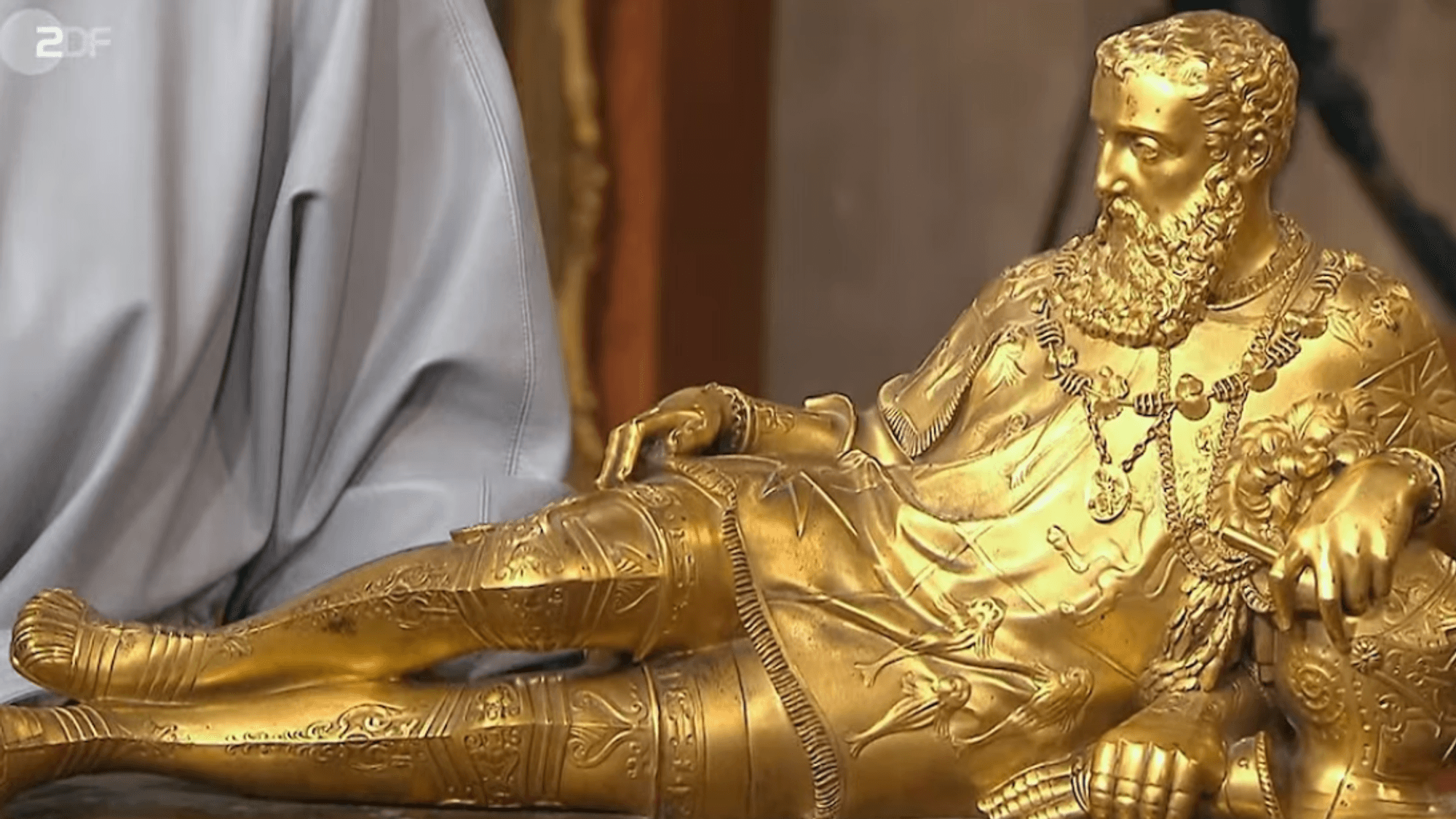 Auf dem Bild liegt der Seemann auf der Seite und blickt nach links. Die Staue ist vergoldet.