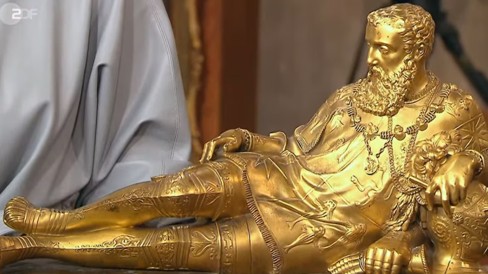 Auf dem Bild liegt der Seemann auf der Seite und blickt nach links. Die Staue ist vergoldet.