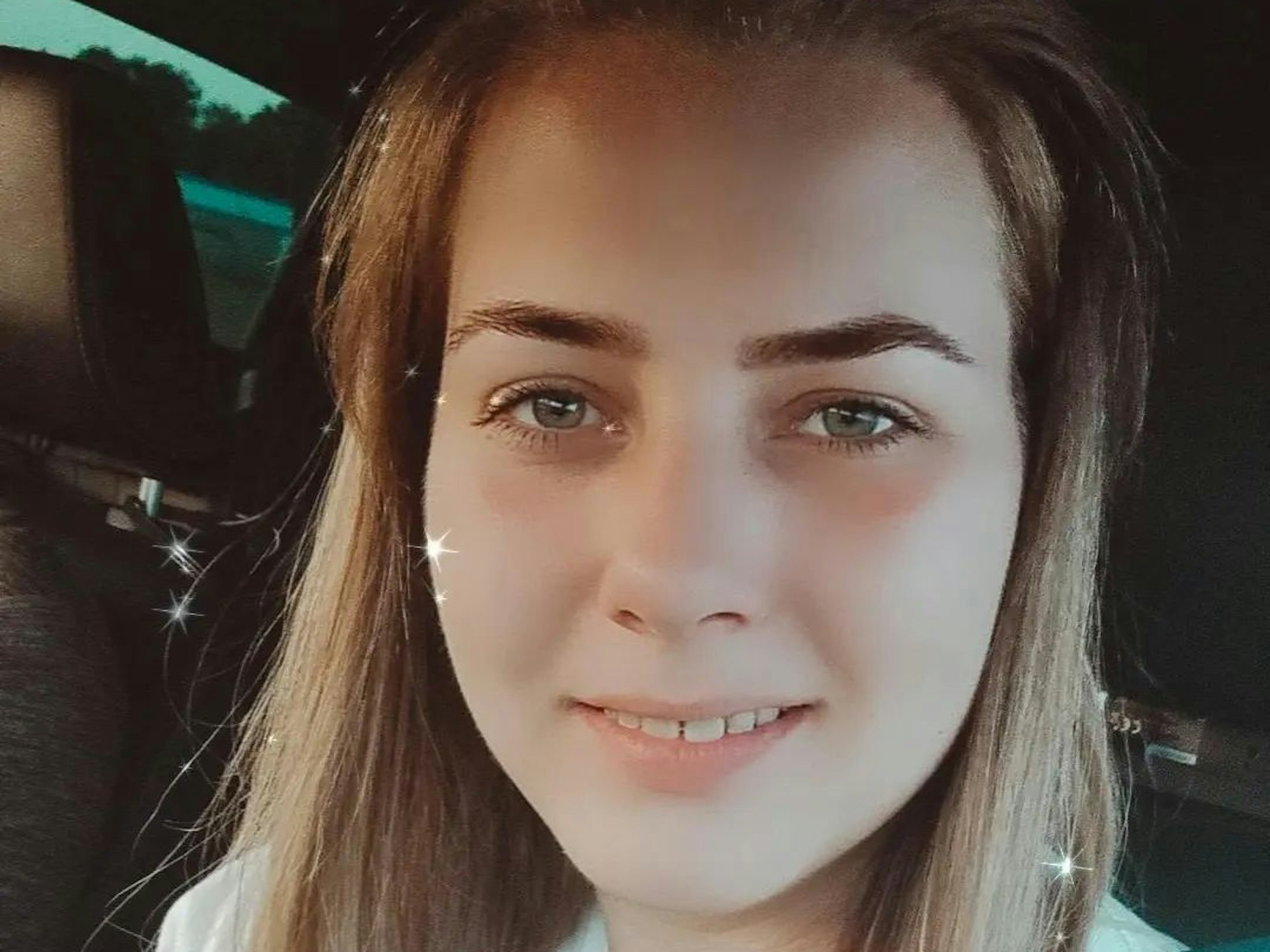 Sarafina posiert für ein Selfie im Auto. Sie trägt ein weißes Shirt und lächelt in die Kamera.