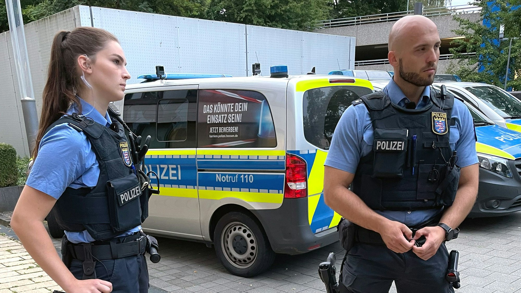 Polizeianwärterin Milena Encarnacao und ihr Ausbilder Dominique Haas stehen in Polizeiuniform vor einem Streifenwagen.