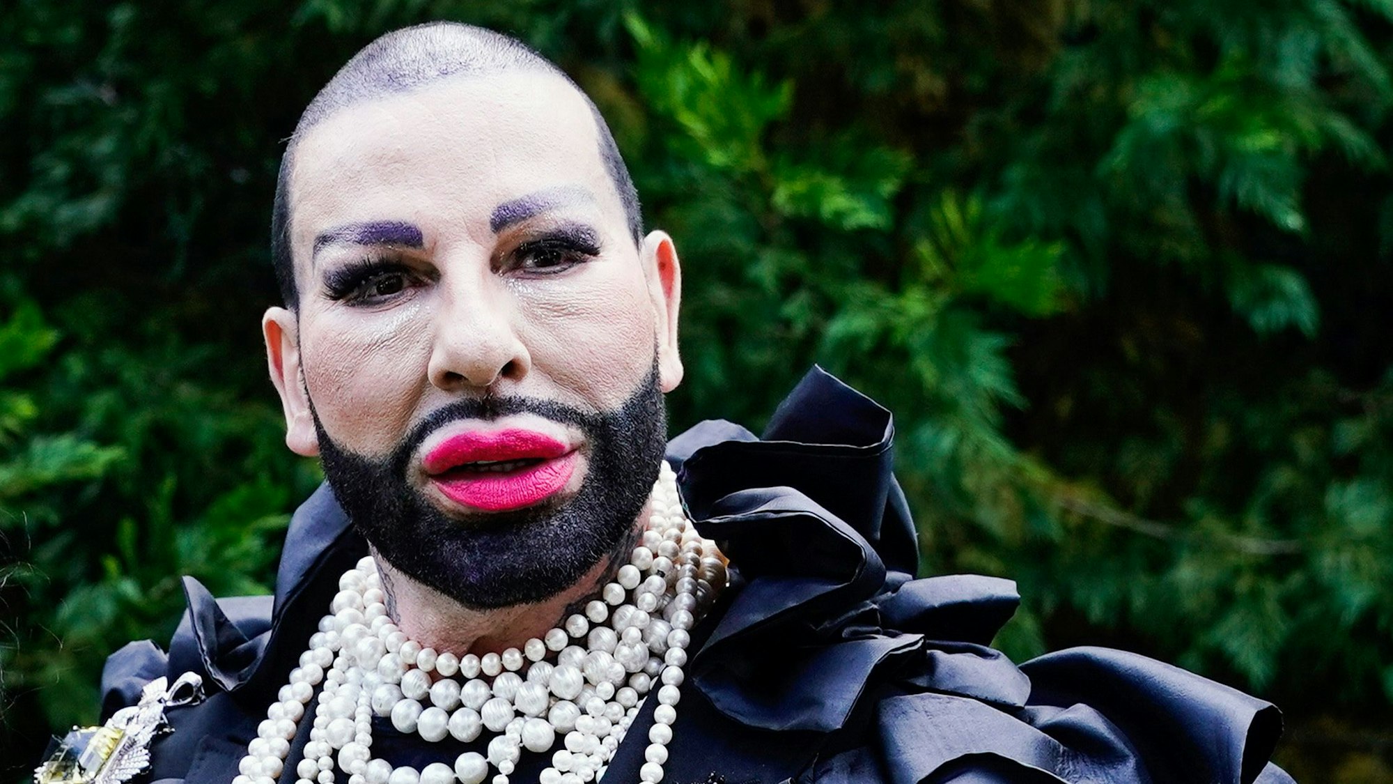 Harald Glööckler posiert in einem schwarzen Hemd und Perlenketten für die Kamera.