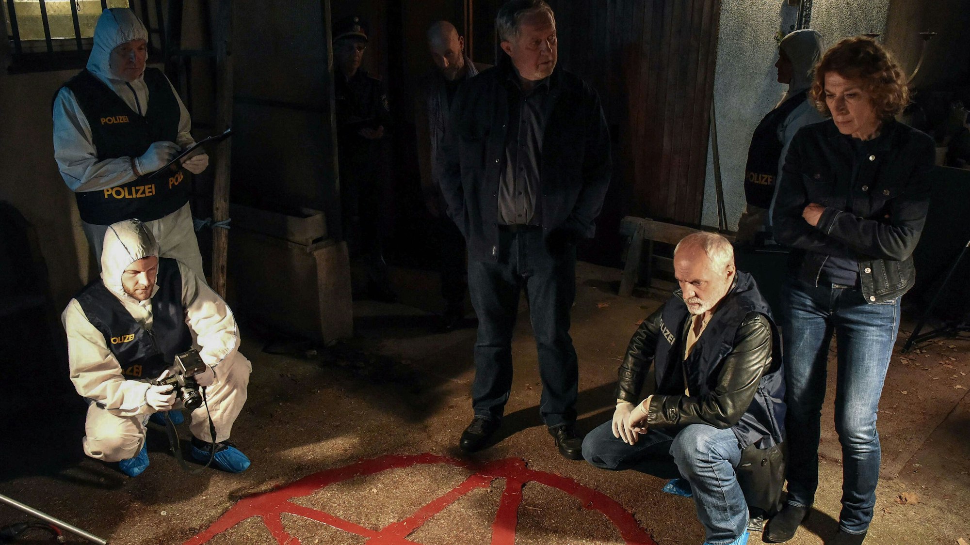 Sechs Menschen stehen um ein rotes Pentagramm, das auf den Boden gemalt ist.