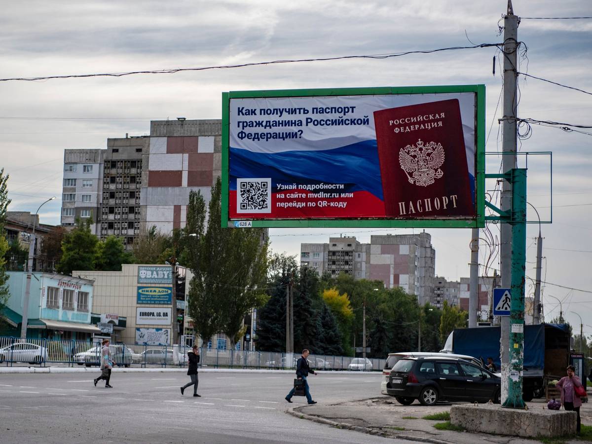 Ukraine, Luhansk: Menschen überqueren eine Straße mit einem Plakat mit der Aufschrift ‚Wie man den Pass eines russischen Bürgers bekommt‘.
