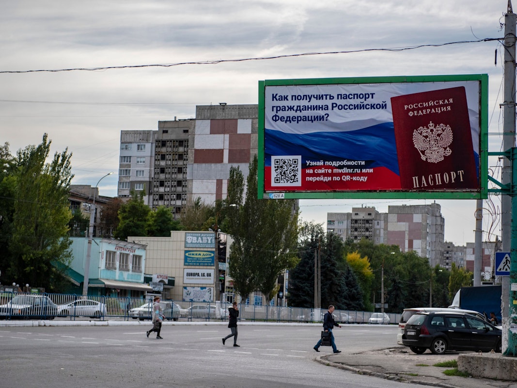 Ukraine, Luhansk: Menschen überqueren eine Straße mit einem Plakat mit der Aufschrift ‚Wie man den Pass eines russischen Bürgers bekommt‘.