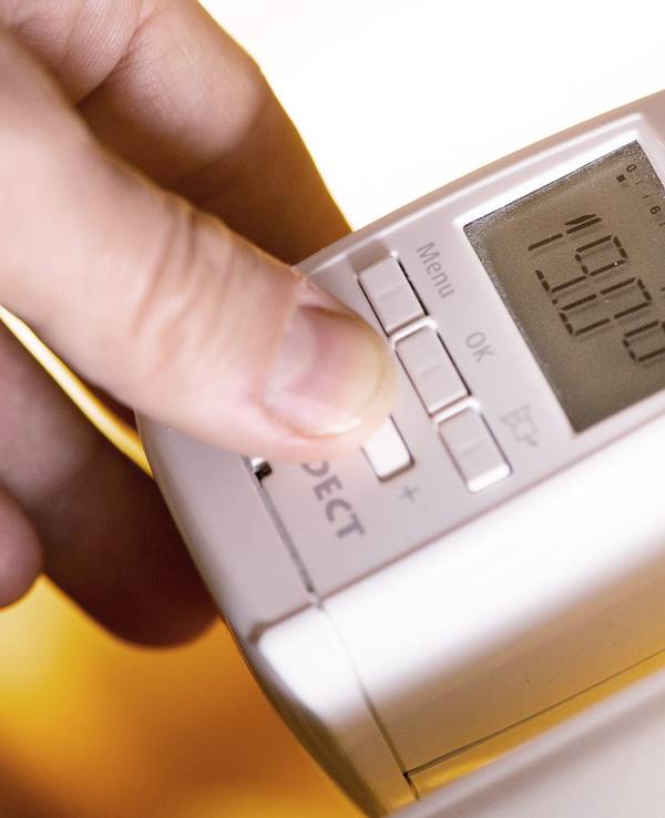 Eine Hand fasst an ein modernes Thermostat mit digitaler Anzeige an einer Heizung, wobei 19. Grad Celsius auf einem kleinen Display angezeigt werden.