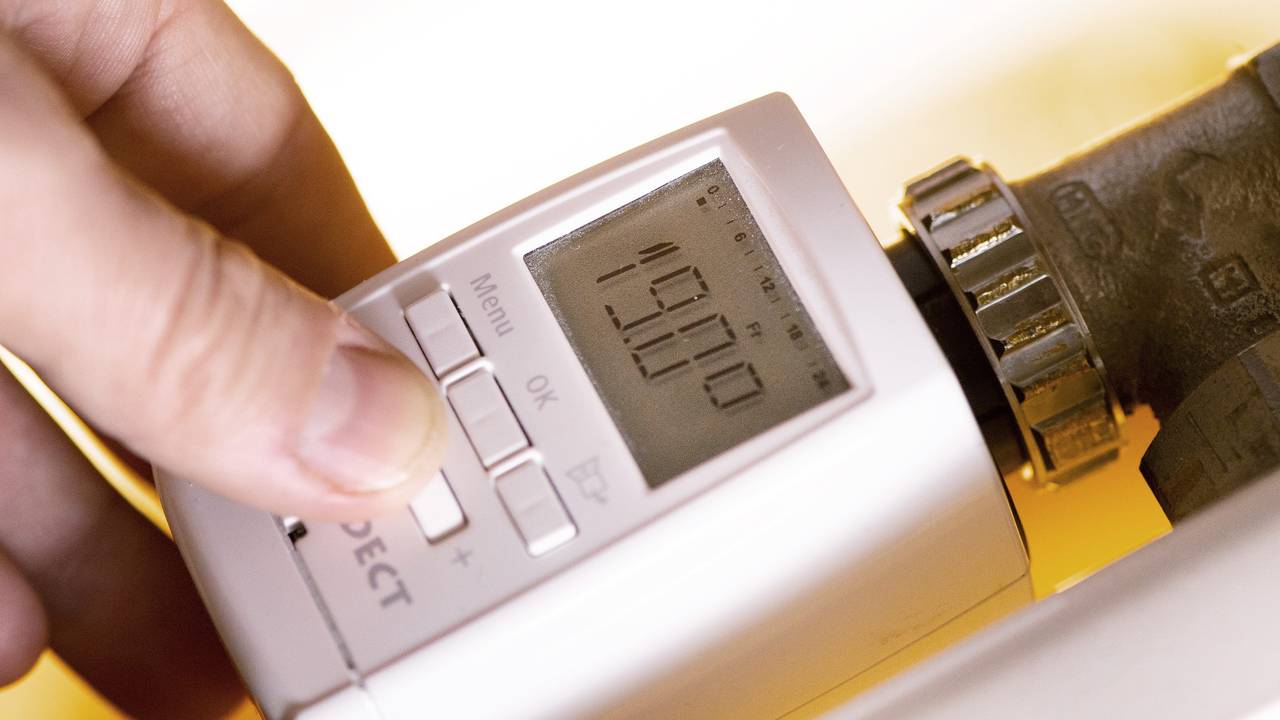 Eine Hand fasst an ein modernes Thermostat mit digitaler Anzeige an einer Heizung, wobei 19. Grad Celsius auf einem kleinen Display angezeigt werden.