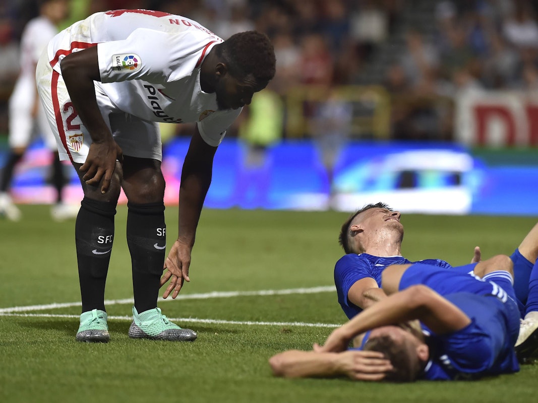 Joris Gnagnon, damals noch im Trikot des FC Sevilla, bückt sich, während zwei Gegenspieler verletzt auf dem Rasen liegen.