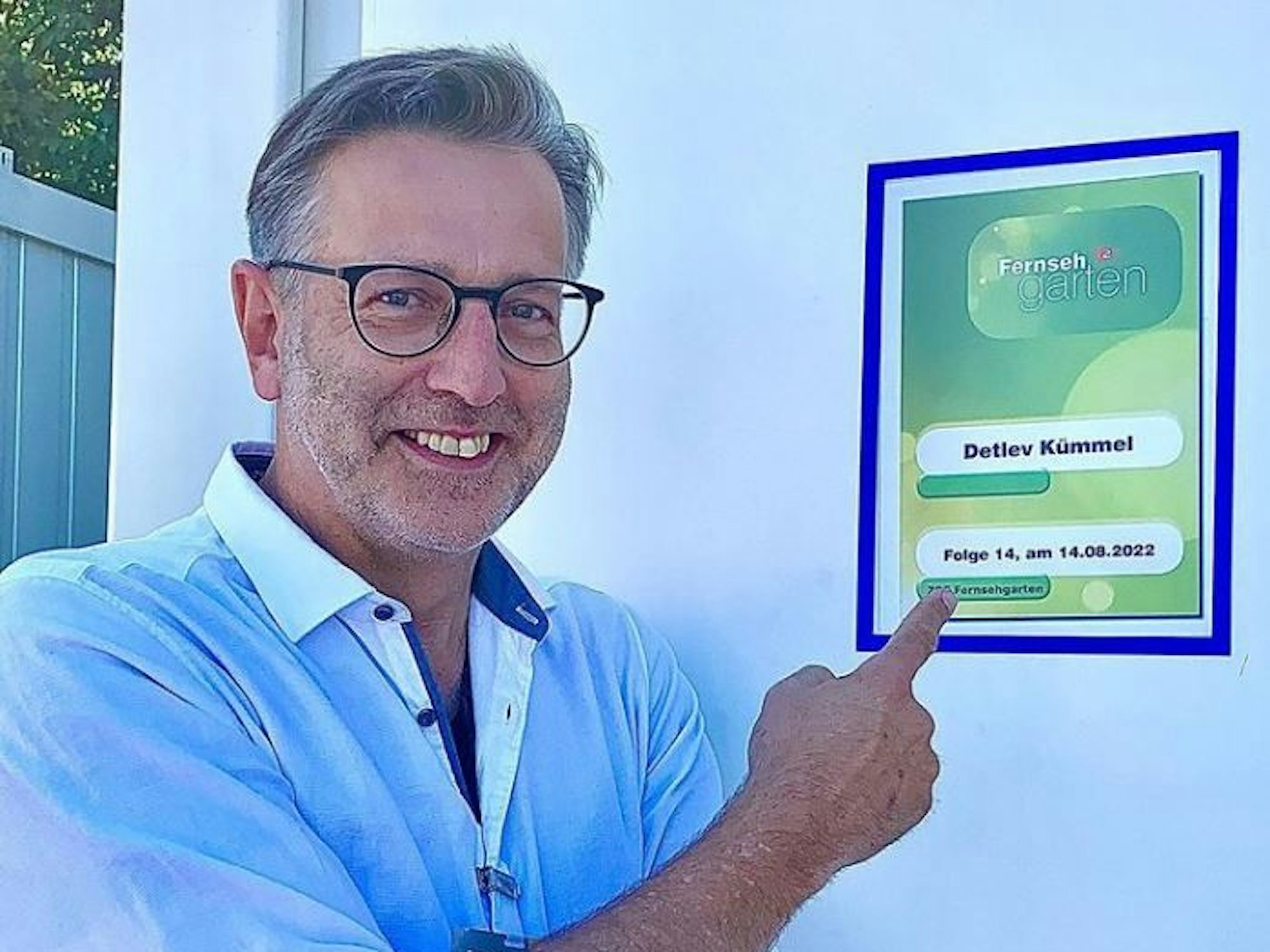 Detlev Kümmel lacht auf einem Foto fröhlich in die Kamera und zeigt auf eine Werbetafel des ZDF-„Fernsehgarten“.