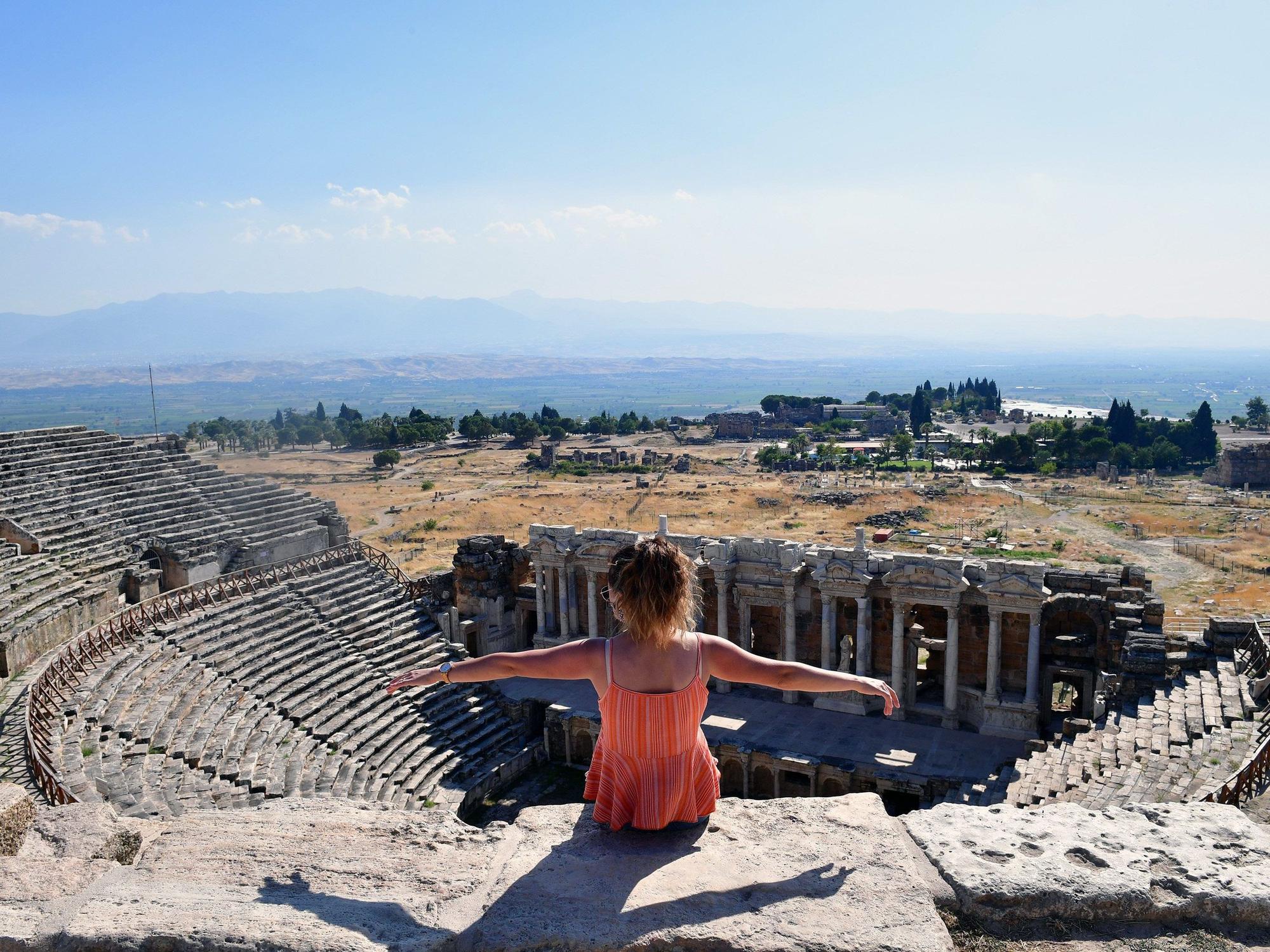 Eine Touristin breitet die Arme aus während sie auf den Ruinen der antiken Stadt und UNESCO-Weltkulturerbe Hierapolis sitzt.