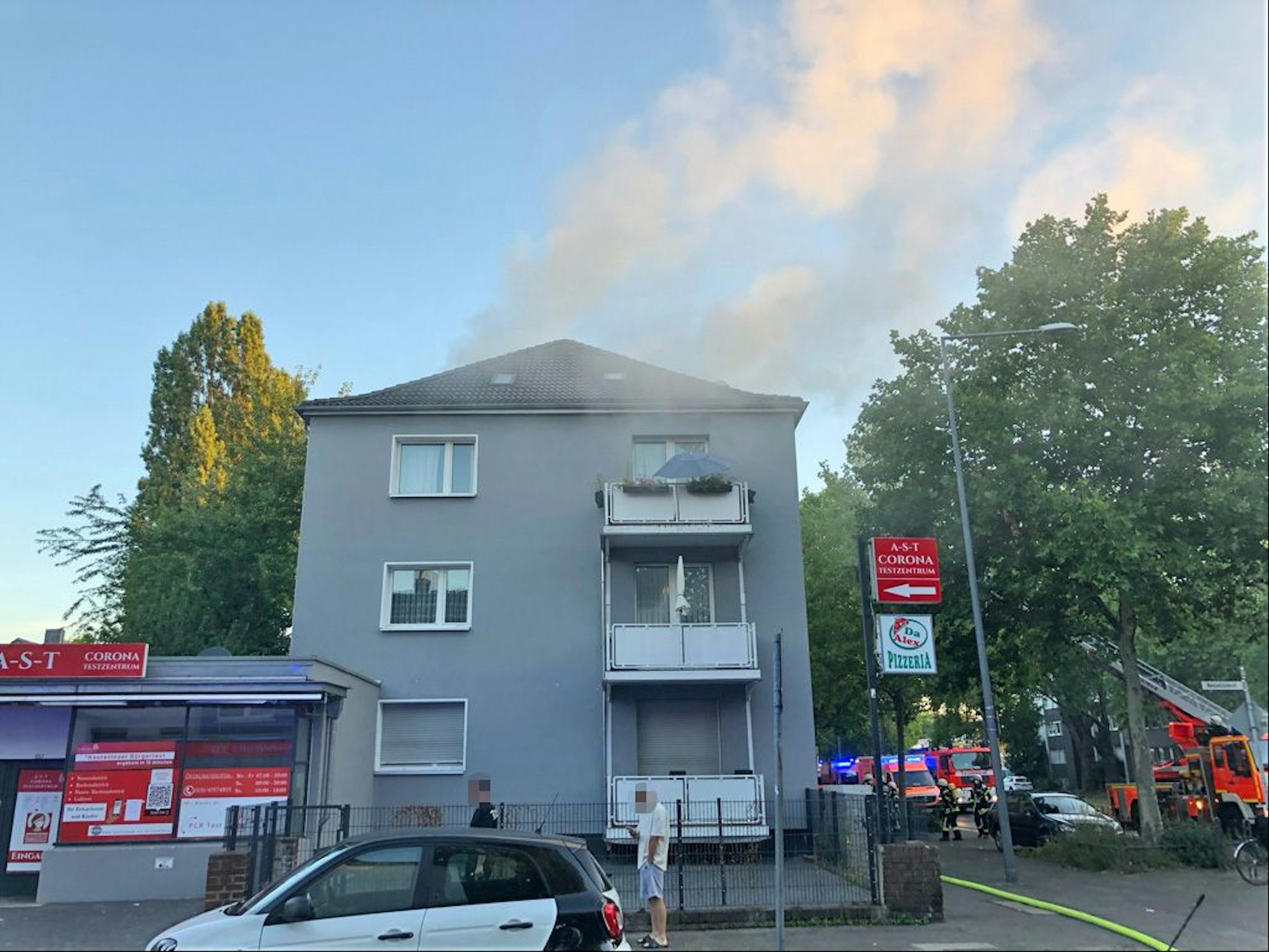 Qualm steigt aus einer Mehrfamilienhaus an einer Straße in Köln. Vor dem Haus steht die Feuerwehr.