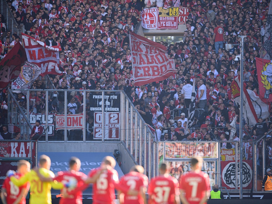 Die FC-Spieler umarmen sich bei einer Schweigeminute, Kölner Fans schwenken Fahnen.