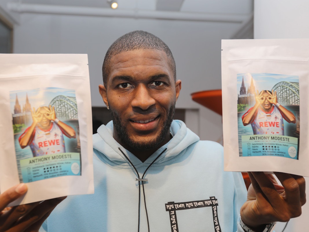 Anthony Modeste hält zwei Tüten des Kaffees in der Hand, auf dem er selbst in Jubelpose zu sehen ist.