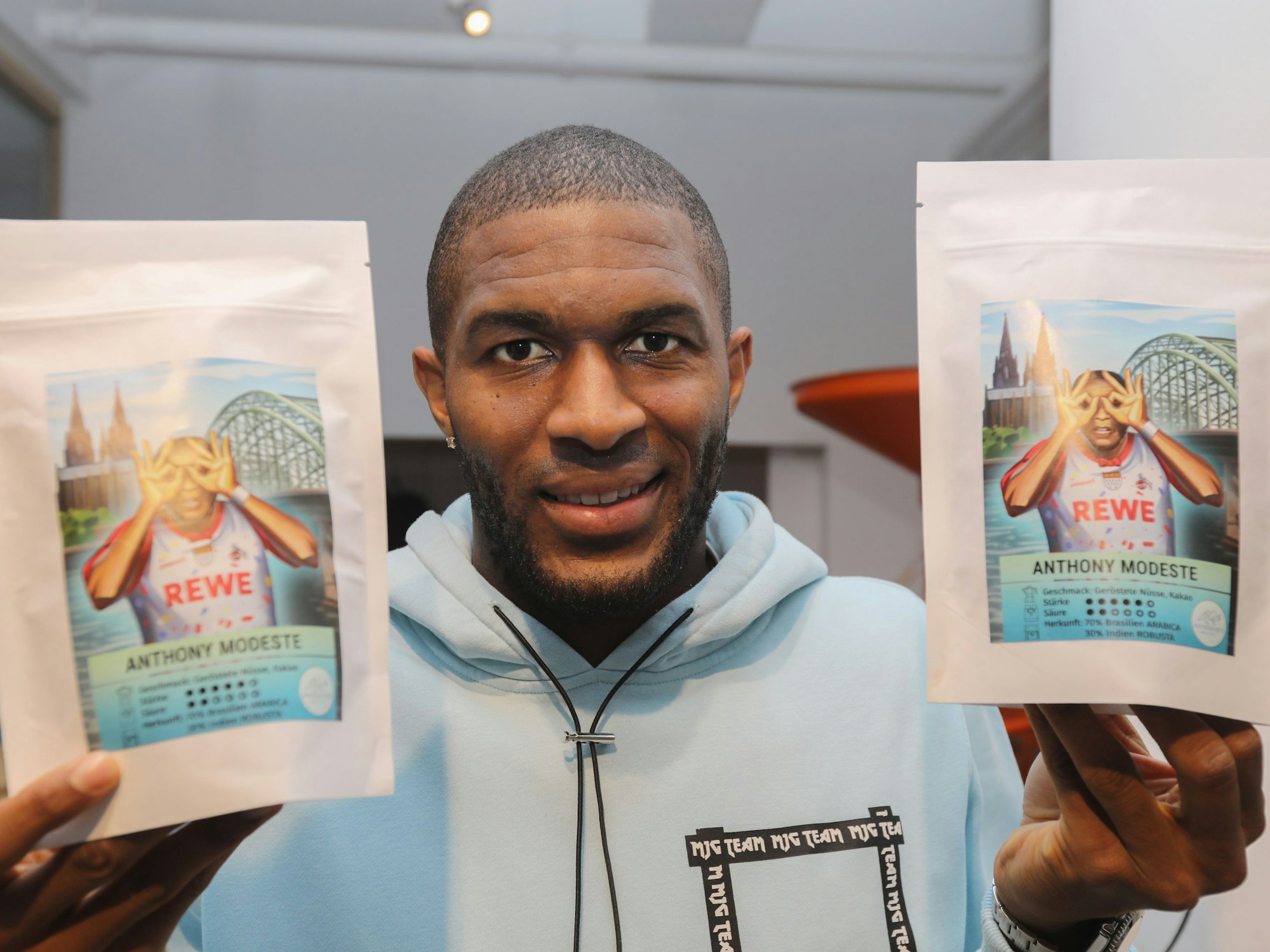 Anthony Modeste hält zwei Tüten des Kaffees in der Hand, auf dem er selbst in Jubelpose zu sehen ist.