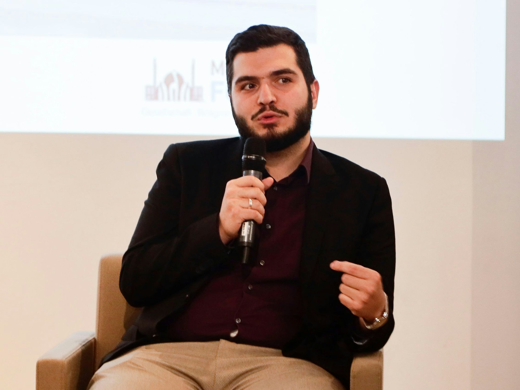 Mustafa Kader, Religionsbeauftragter der Ditib, sitzt in einem Stuhl und spricht in ein Mikrofon. Er setzt den Muezzin-Ruf in Köln ab.