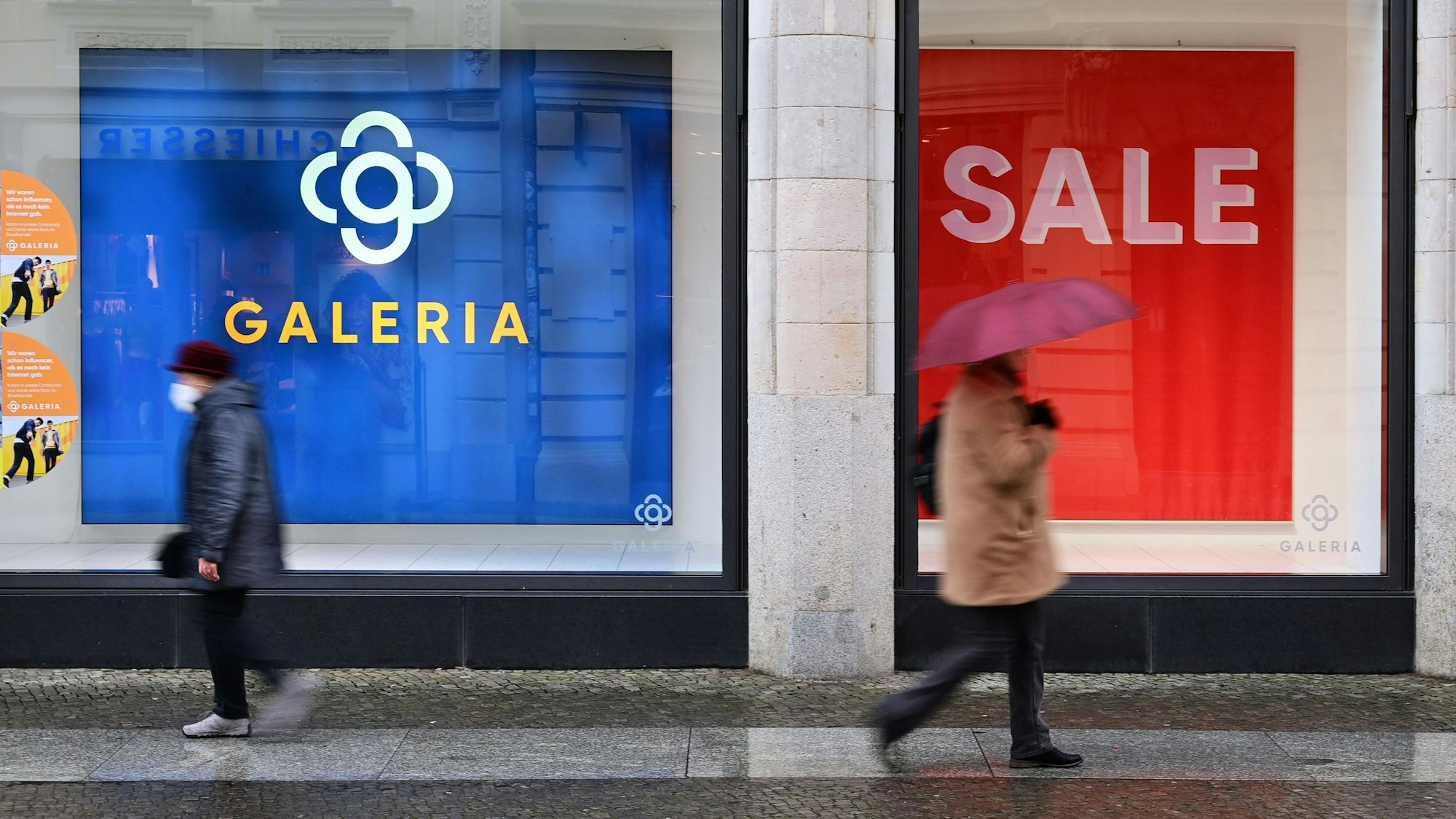 Das undatierte Symbolfoto zeigt zwei Menschen, die an einem Schaufenster mit großen Plakaten vorbeigehen.