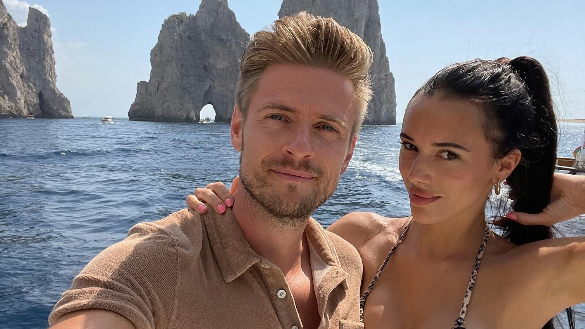 Jörn und Hanna posieren für ein Selfie auf einem Boot. Sie trägt einen Bikini, er ein braunes Poloshirt.