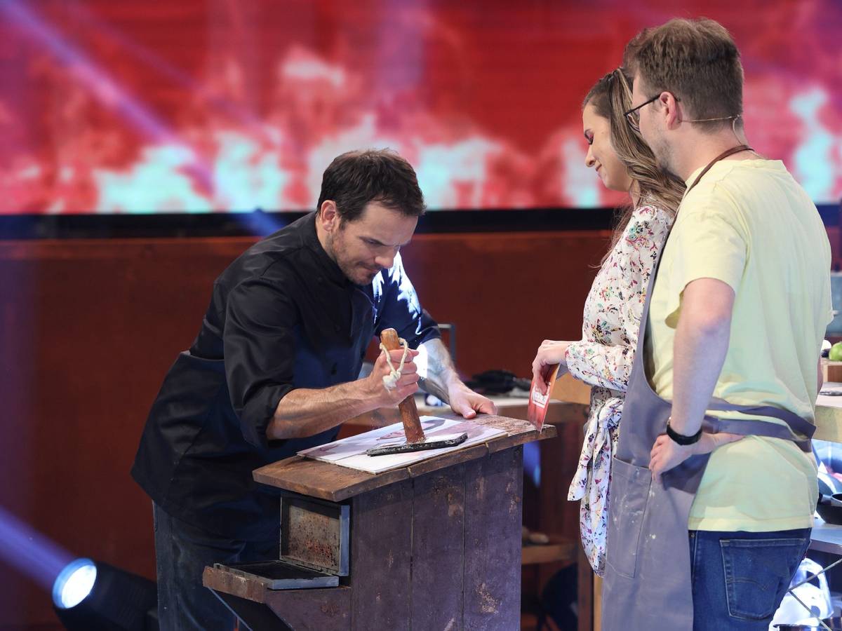 Steffen Henssler demonstriert Laura Wontorra und Maxi Gstettenbauer, wie er etwas an einem Tisch zerstampft.