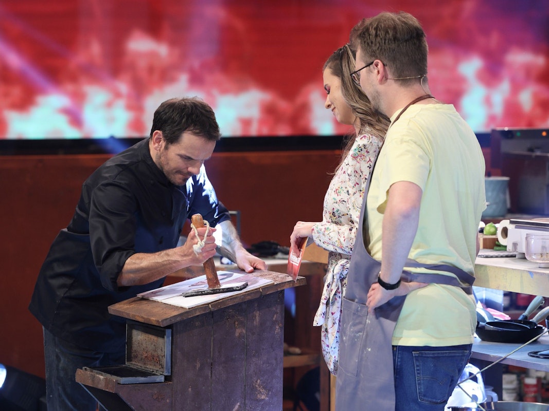 Steffen Henssler demonstriert Laura Wontorra und Maxi Gstettenbauer, wie er etwas an einem Tisch zerstampft.