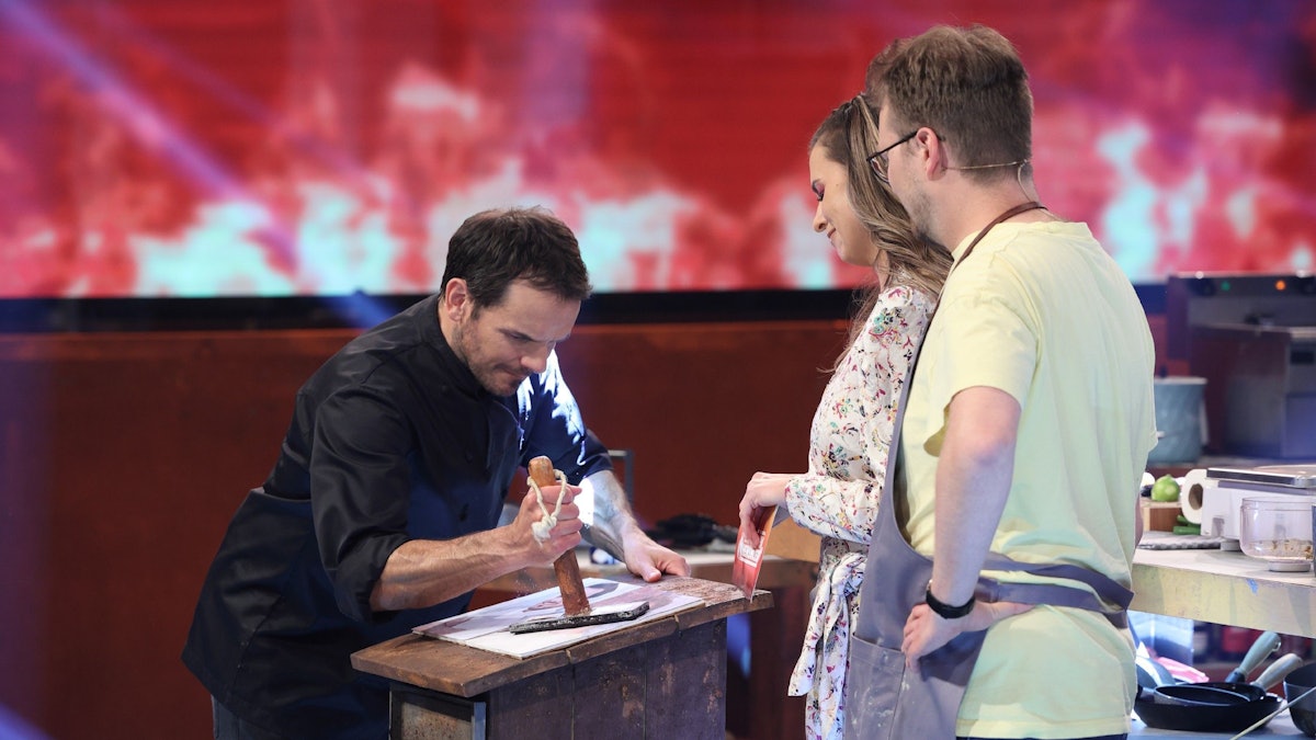 Steffen Henssler demonstriert Laura Wontorra und Maxi Gstettenbauer, wie er etwas an einem Tisch zerstampft.