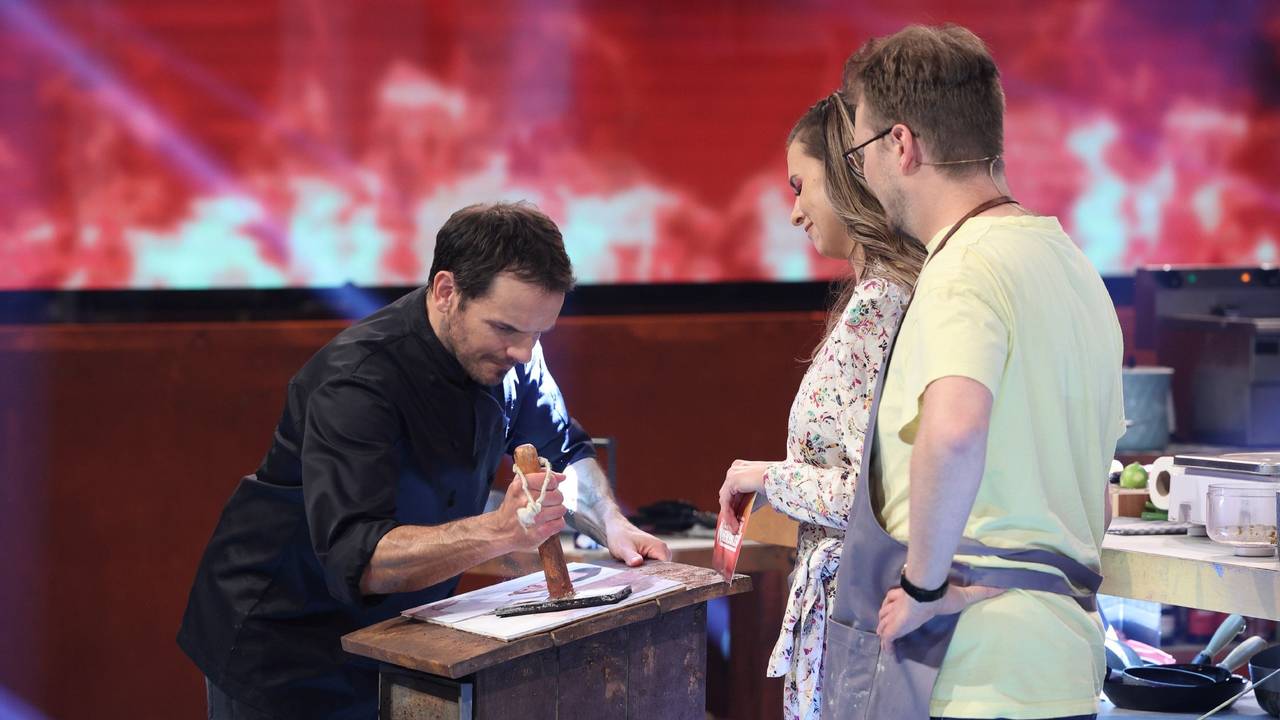 Steffen Henssler demonstriert Laura Wontorra und Maxi Gstettenbauer, wie er etwas an einem Tisch zerstampft.