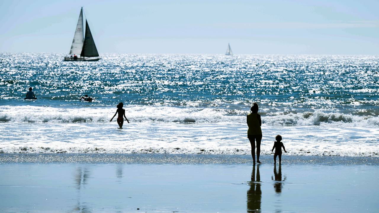 Das undatierte Symbolfoto zeigt einen Strand und das dahinterliegende Meer mit mehreren Menschen und zwei Segelbooten.
