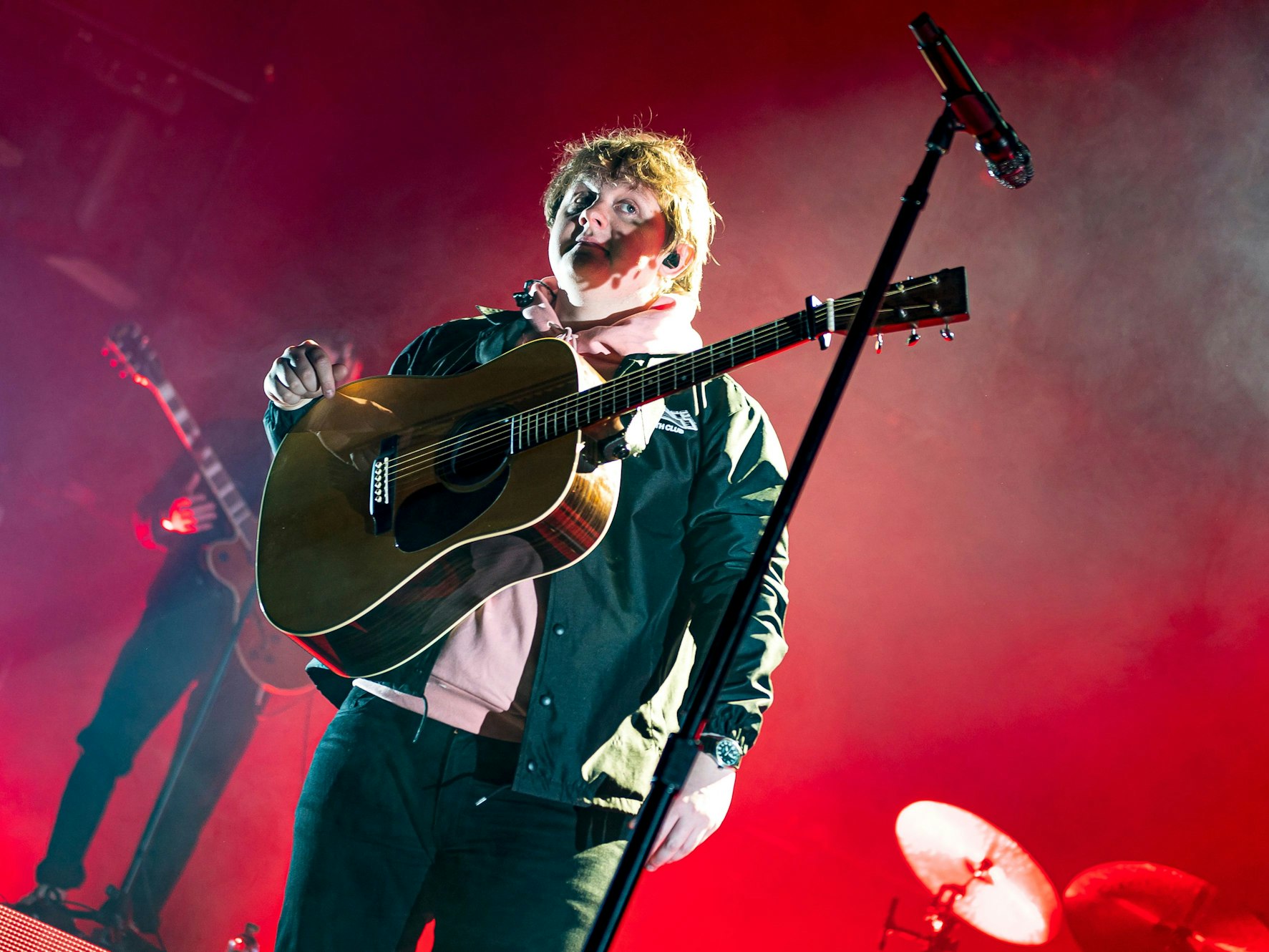 Lewis Capaldi vor einem roten Hintergrund und mit Gitarre bei einem Konzert in Köln.
