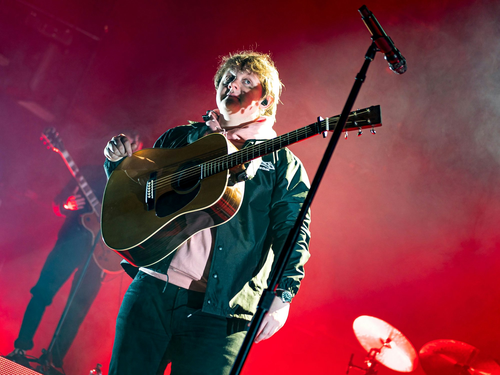 Lewis Capaldi vor einem roten Hintergrund und mit Gitarre bei einem Konzert in Köln.