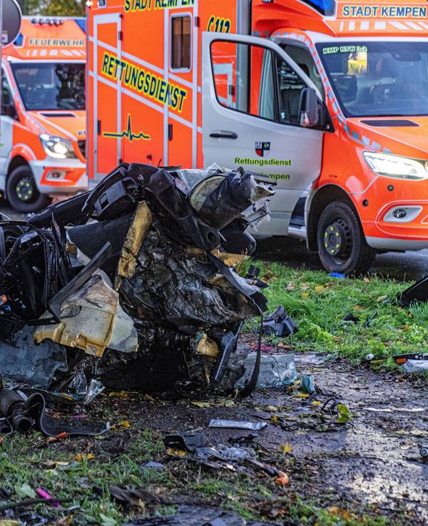 Das Wrack eines Autos liegt völlig zerfetzt neben einem Baum. Daher stehen zwei Rettungswagen auf der Straße.