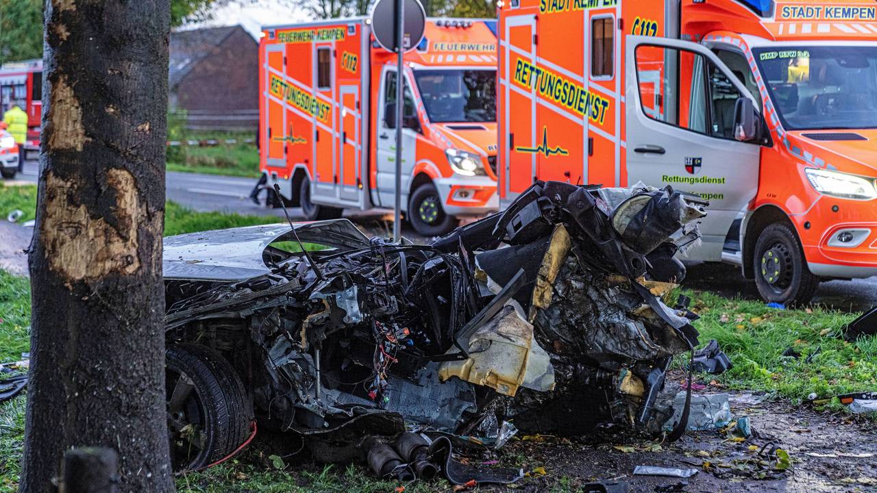 Das Wrack eines Autos liegt völlig zerfetzt neben einem Baum. Daher stehen zwei Rettungswagen auf der Straße.