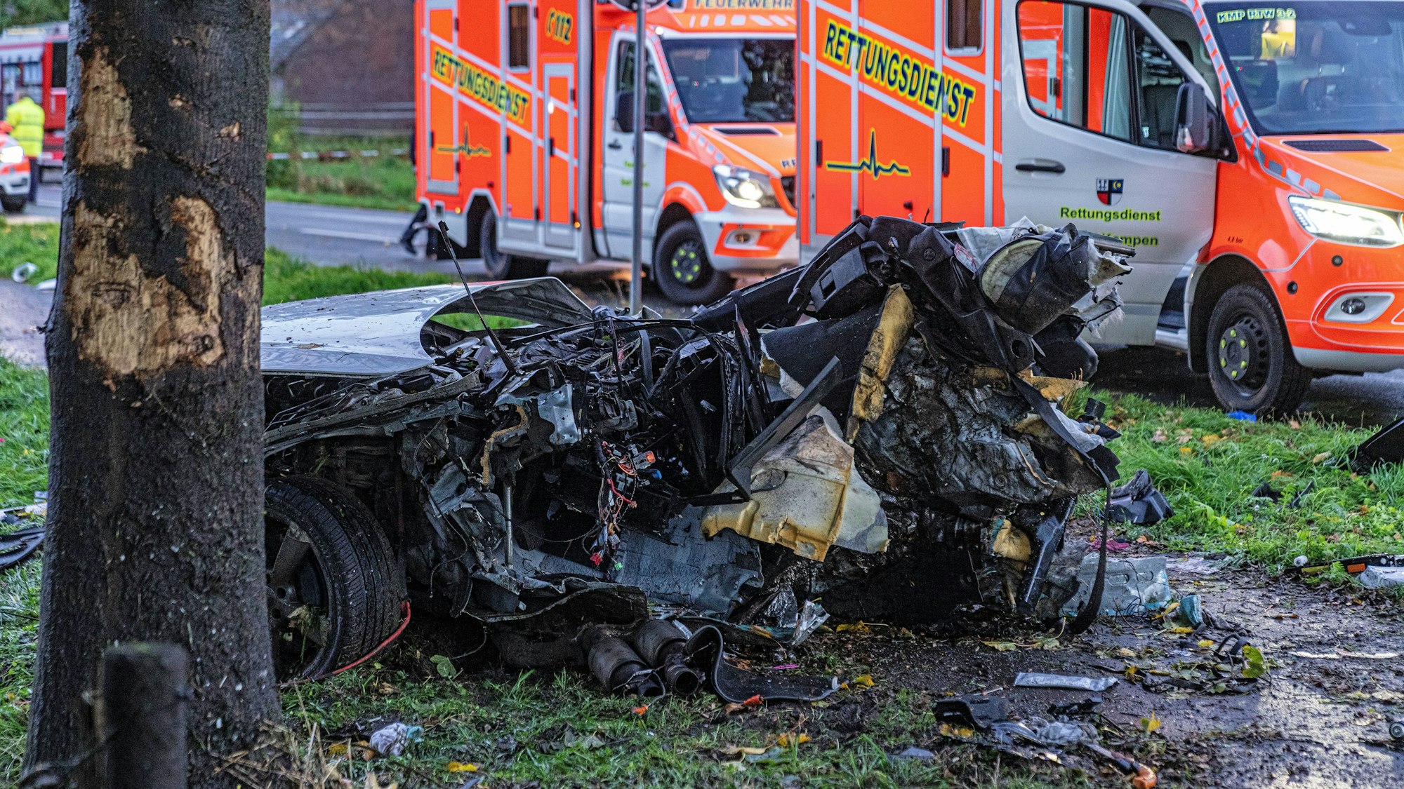 Das Wrack eines Autos liegt völlig zerfetzt neben einem Baum. Daher stehen zwei Rettungswagen auf der Straße.