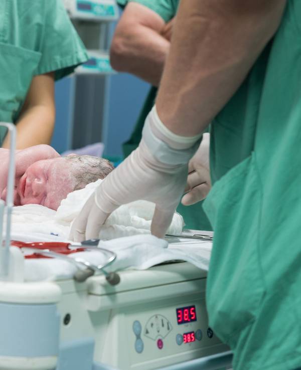 Das Symbolfoto aus dem Jahr 2016 zeigt ein Neugeborenes, das von Krankenhauspersonal in grünen Kitteln umgeben ist.