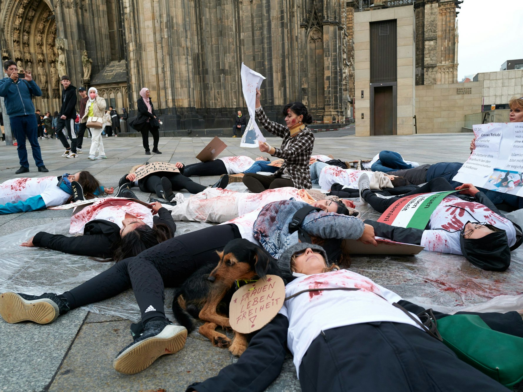 Rund 20 Personen (überwiegend Frauen) liegen in Köln auf der Domplatte – ihre T-Shirts sind mit Kunstblut beschmiert.