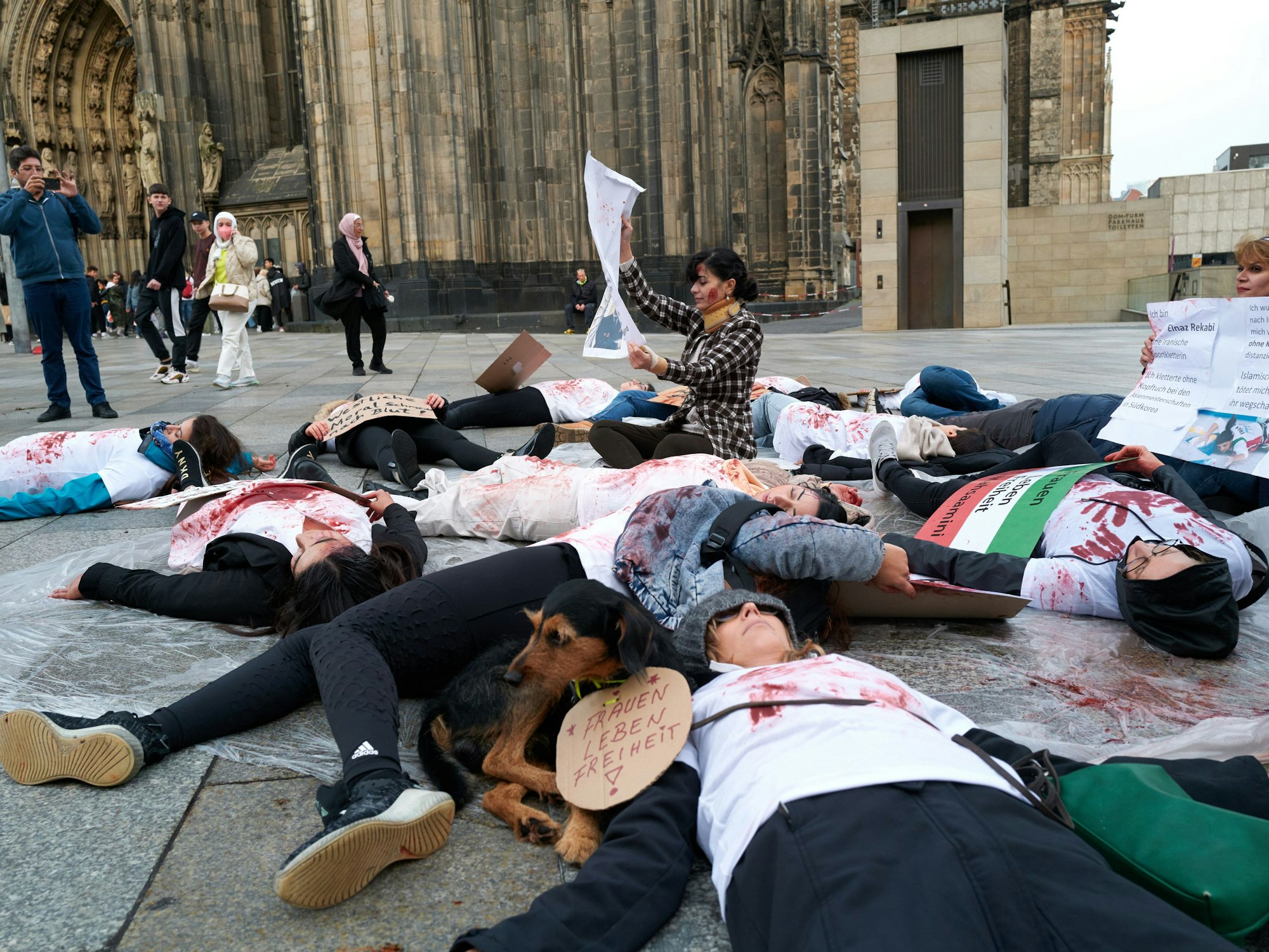 Rund 20 Personen (überwiegend Frauen) liegen in Köln auf der Domplatte – ihre T-Shirts sind mit Kunstblut beschmiert.