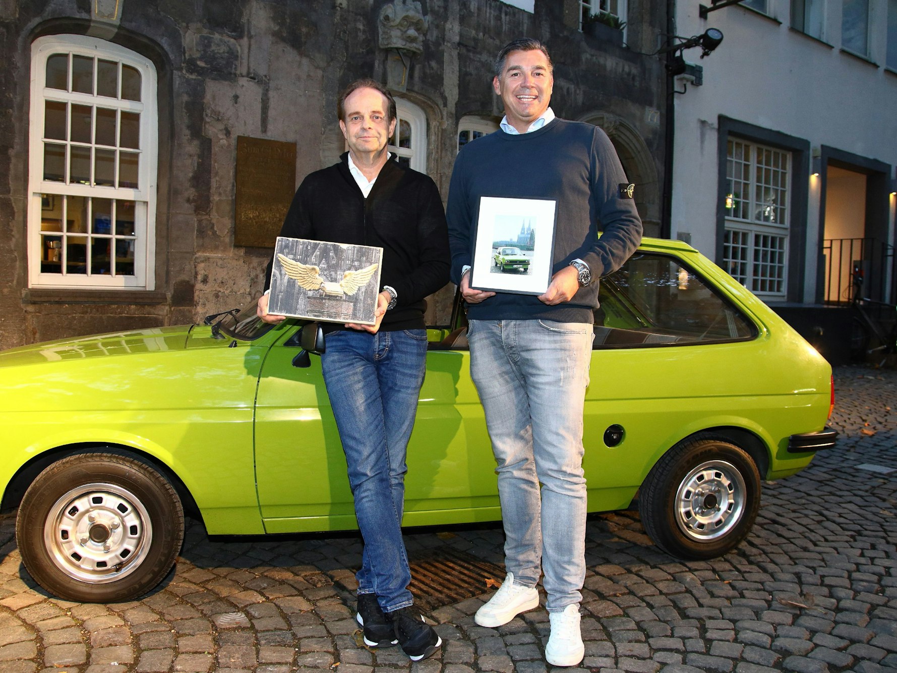 Oliver Wunderlich und Geert Dilien stehen mit Fotos vor einem alten Ford Fiesta in Köln.