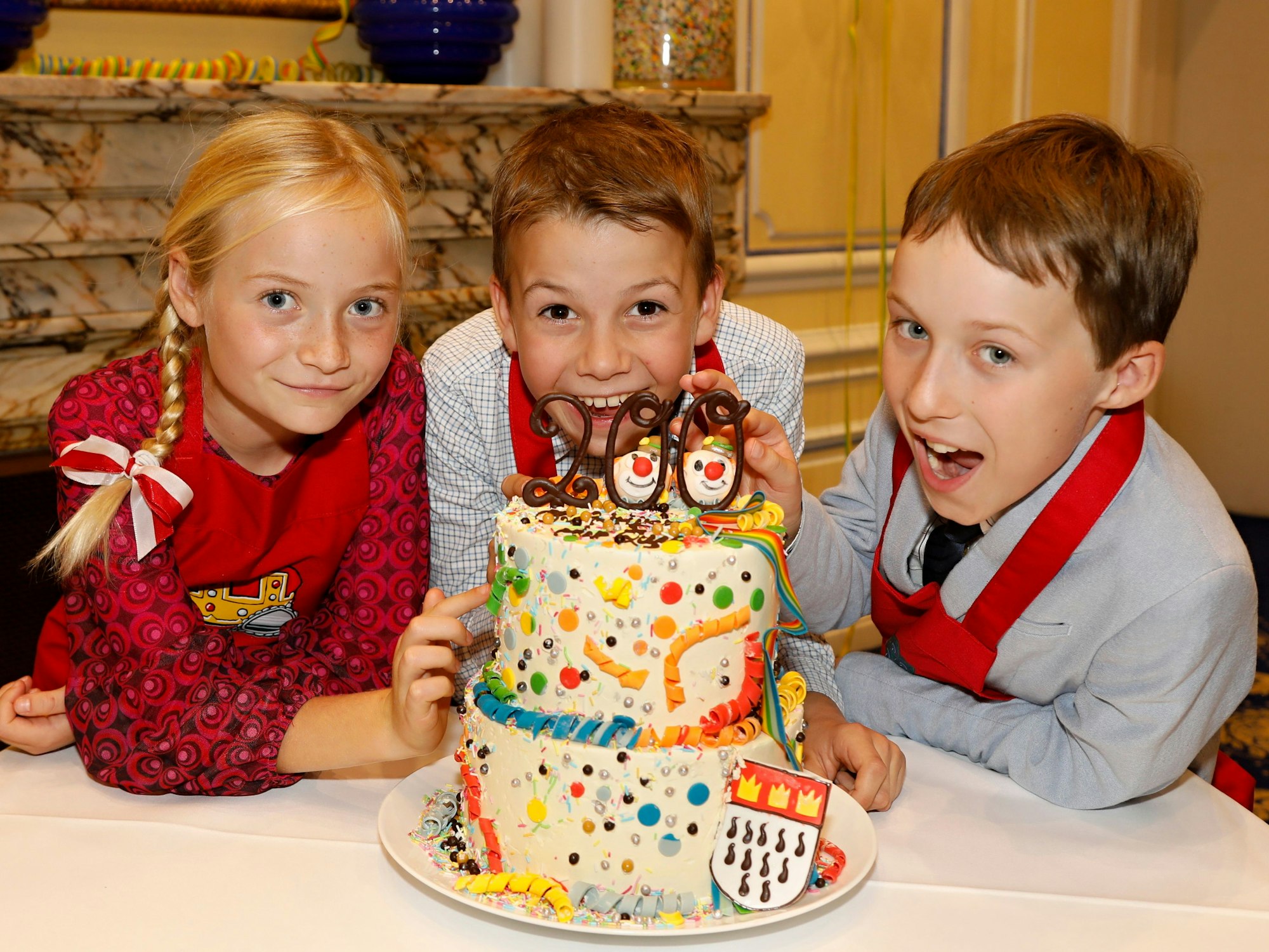 Das neue Kölner Kinderdreigestirn vor einer Torte zum 200-jährigen Jubiläum des Festkomitees.