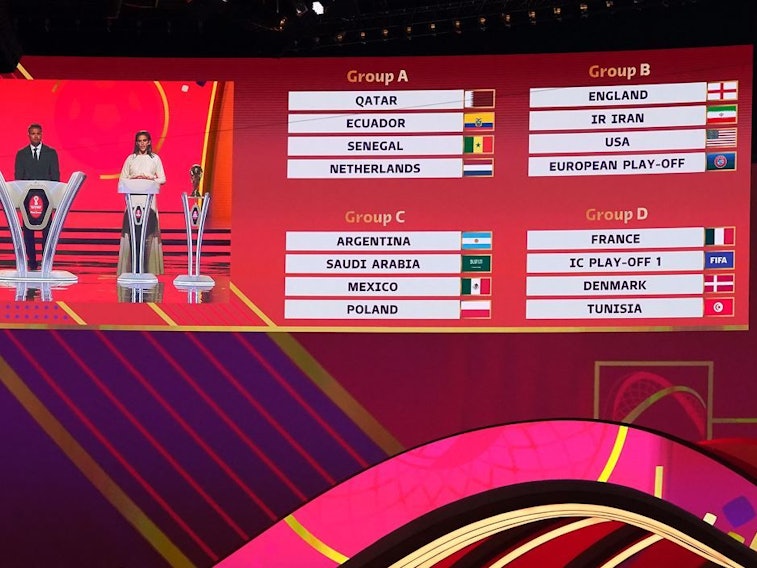 WM-Auslosung der Vorrunde in Doha. Die Auslosungsleiter Sam Johnson, Jermaine Jenas und Carli Lloyd sind auf einer Anzeige neben den Gruppen A,B,C und D zu sehen.