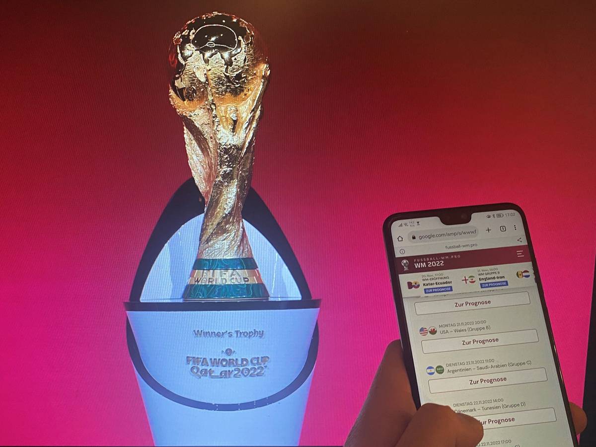 Ein Handy mit einem geöffneten Tippspiel, im Hintergrund der Pokal der Fußball-WM.