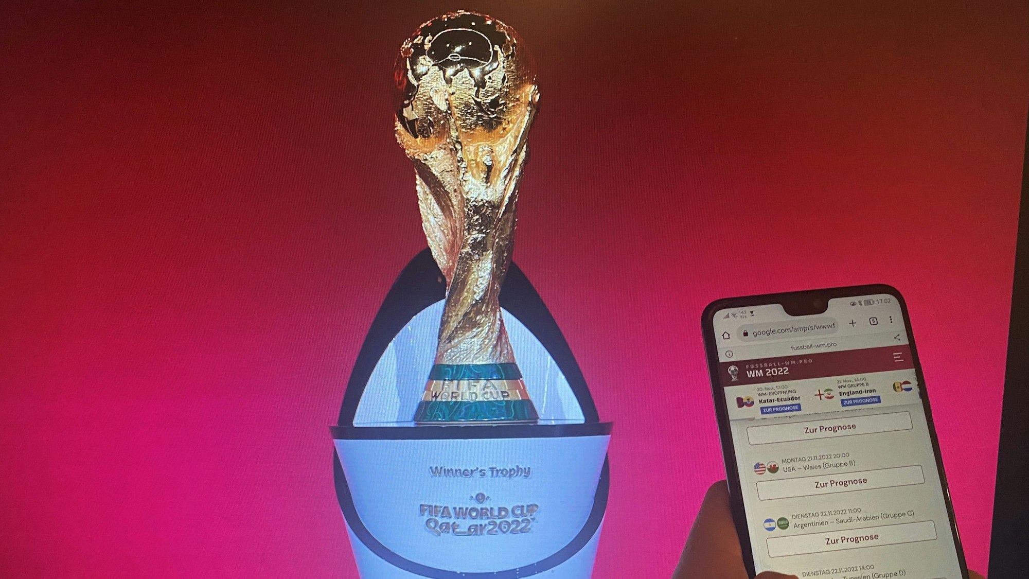 Ein Handy mit einem geöffneten Tippspiel, im Hintergrund der Pokal der Fußball-WM.